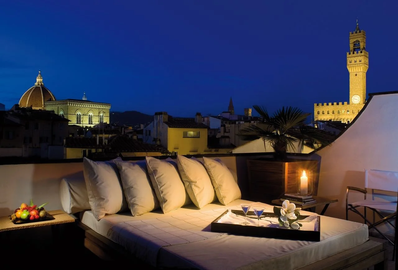 Rooftop balcony at night with a cushioned sofa, tray with drinks and flowers, candle, fruit, palm plant, city skyline with historic buildings and clock tower in Florence, Italy, illuminated