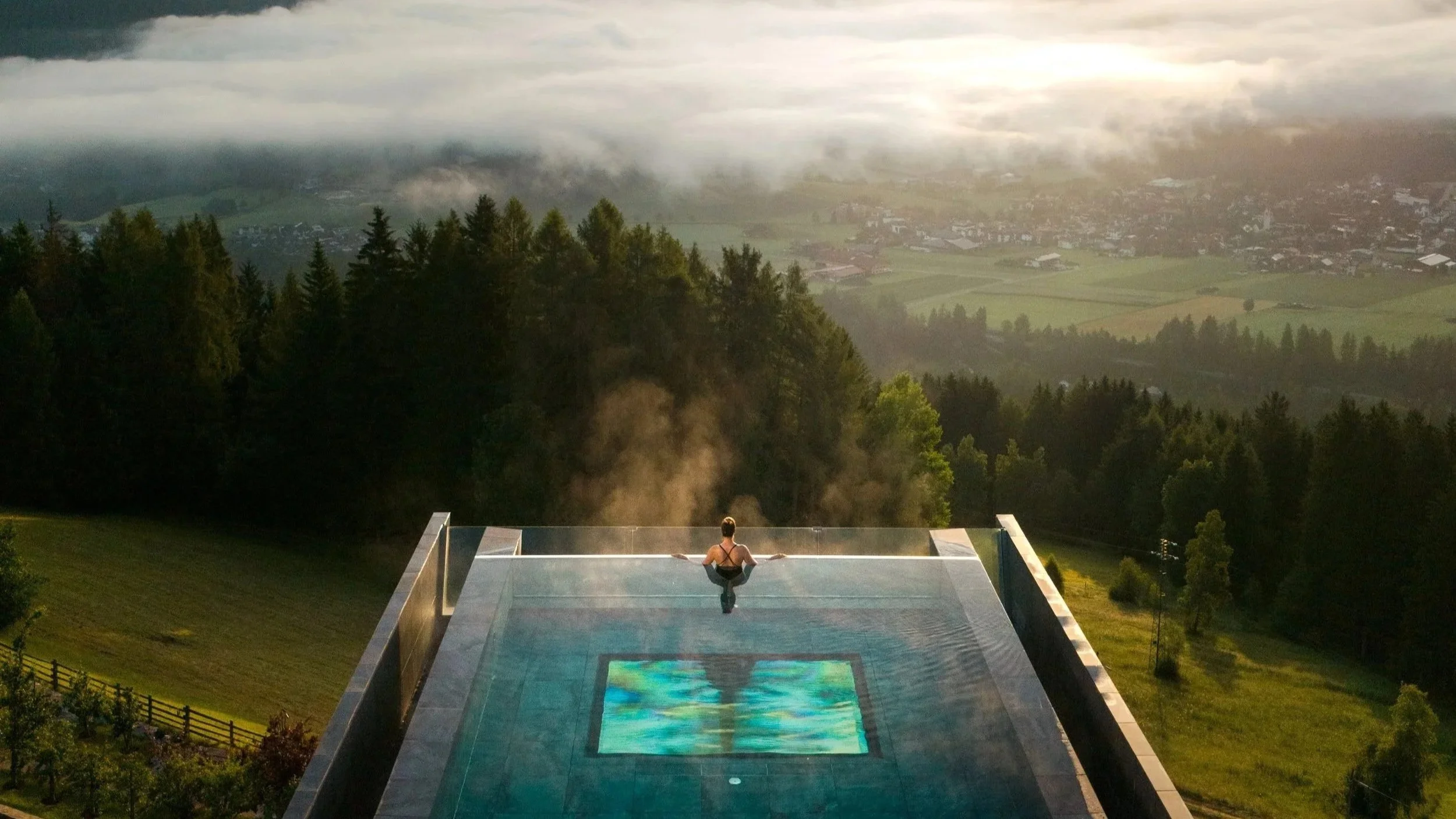A woman sitting on the edge of a floating infinity pool overlooking a scenic landscape of trees, hills, and a valley shrouded in mist and clouds during sunrise or sunset.