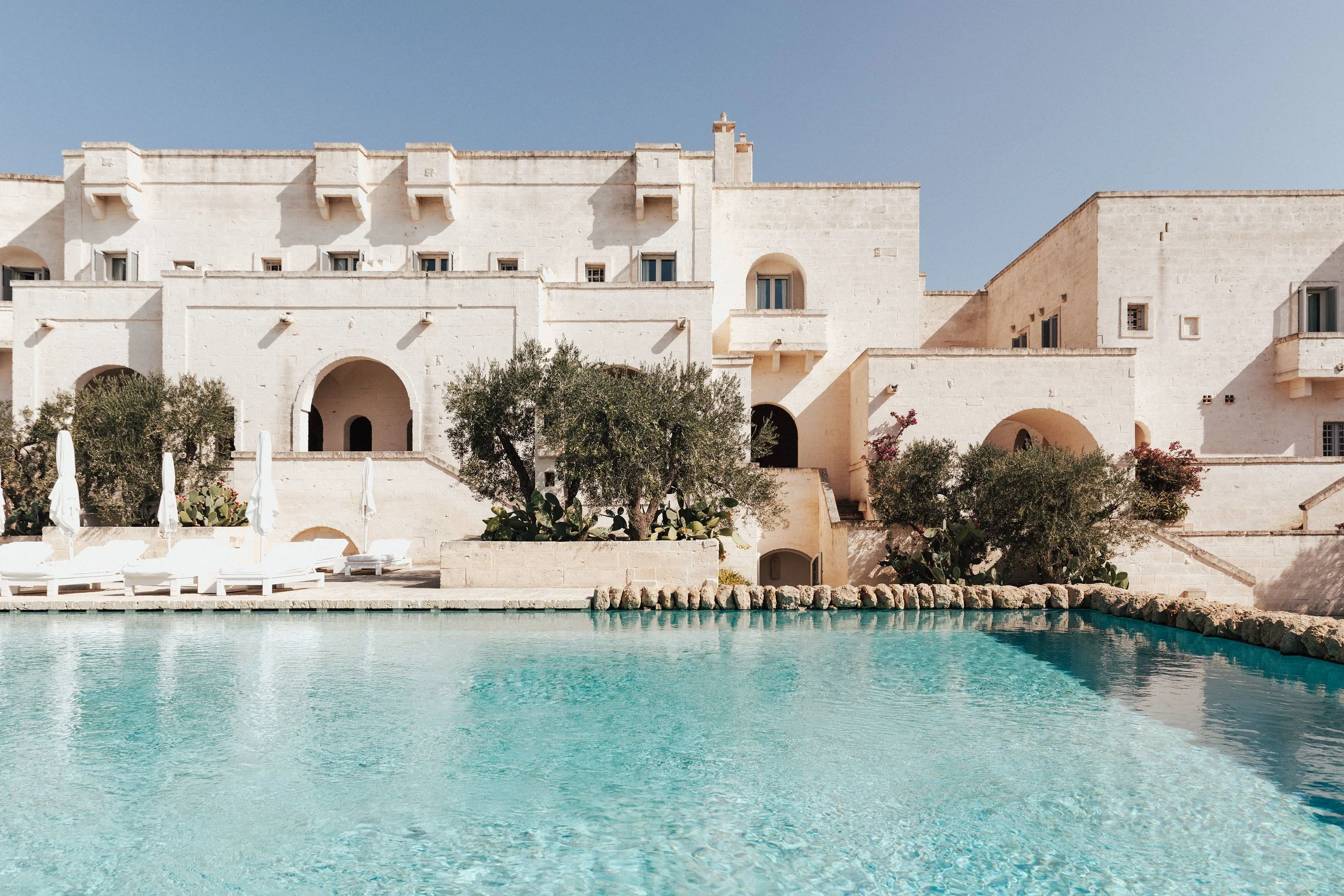 A large white Mediterranean-style building with arched doorways and small windows, surrounded by trees and bushes, overlooking a swimming pool with clear blue water.