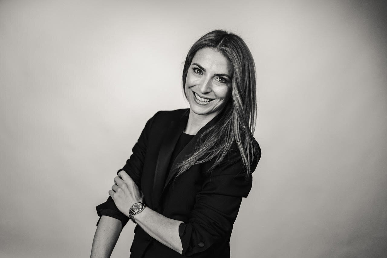 Black and white portrait of a woman with long hair, smiling, wearing a blazer, arms crossed, against a plain background.