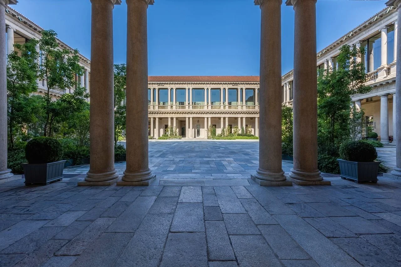 View of a classical building courtyard framed by large stone columns, with a paved walkway and lush green plants on either side.