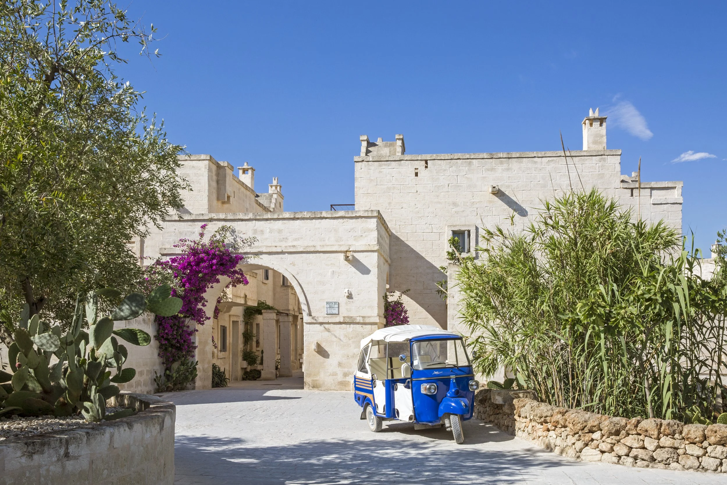 Photo of a historic European town at sunset with limestone buildings, bell towers, and a clock tower with a blue clock face, overlooking an expansive landscape and distant sea.