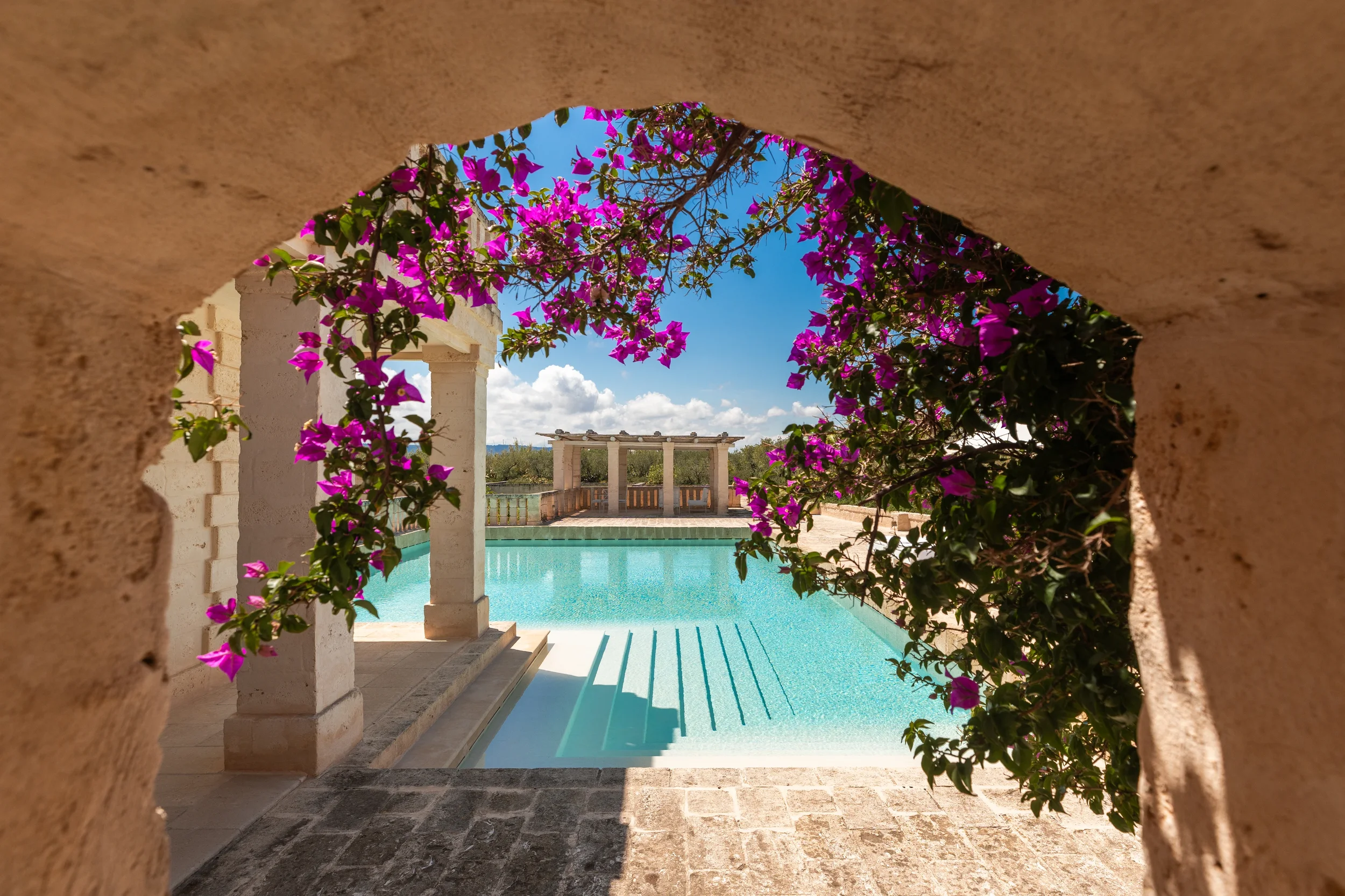 Looking through a small opening with pink bougainvillea flowers over a swimming pool and a white gazebo structure with columns in the background under a blue sky with clouds.