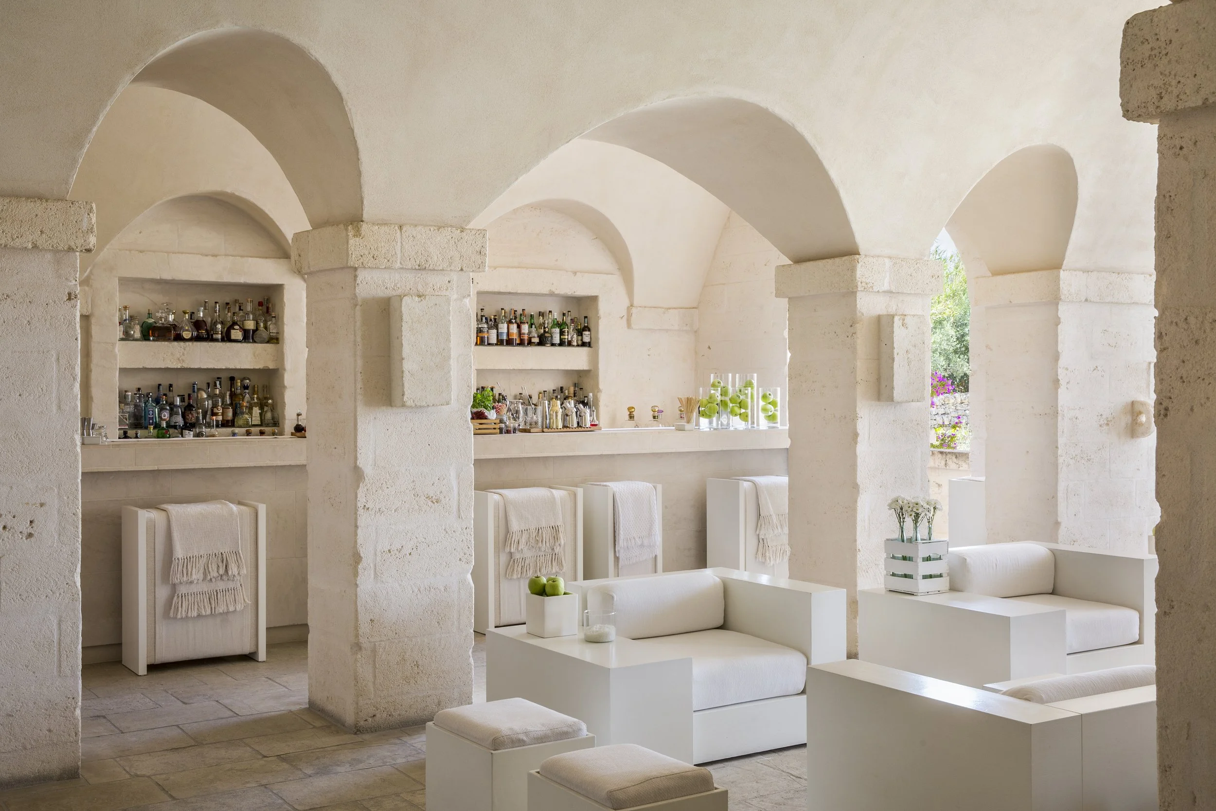 A bright, elegant lounge with white seating and decorative arches, featuring a bar area with numerous bottles, glassware, and green apples on display, and a window revealing greenery outside.