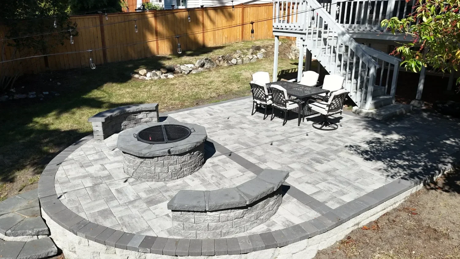 Backyard paver patio installation in Seattle, WA with a circular fire pit, curved seat walls, and a dining area under string lights.