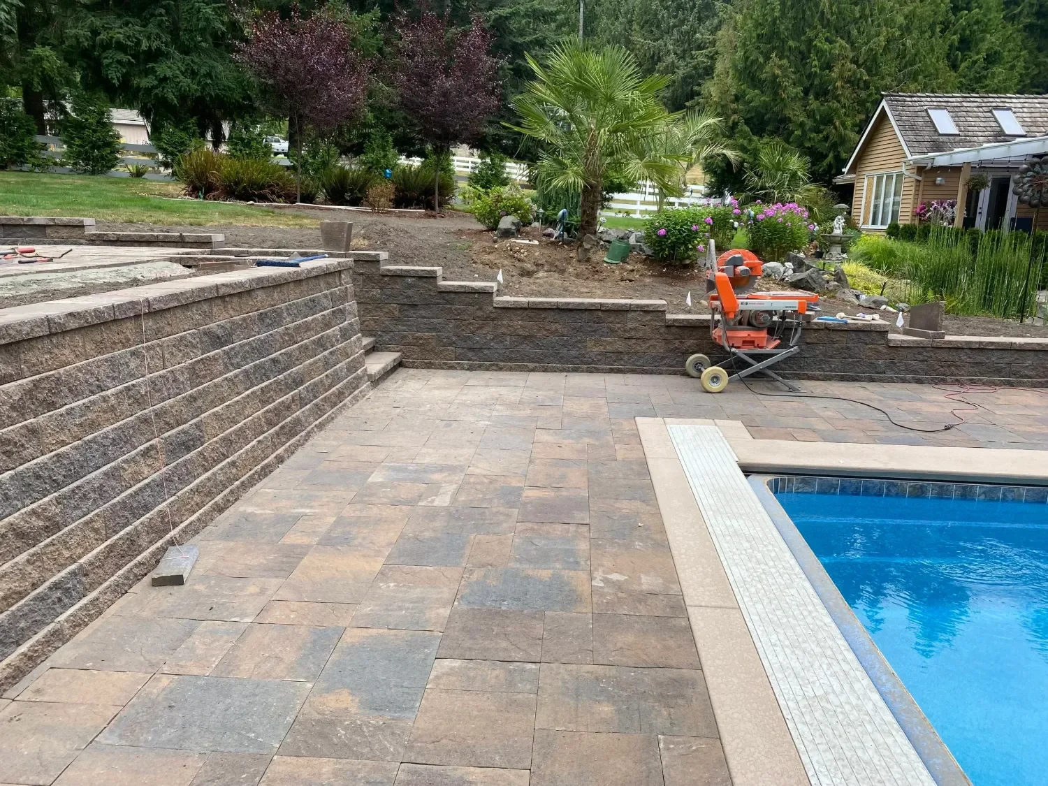 Earth-toned paver pool deck and tiered retaining wall installation in Kirkland, WA with step access and an in-progress hardscape build.