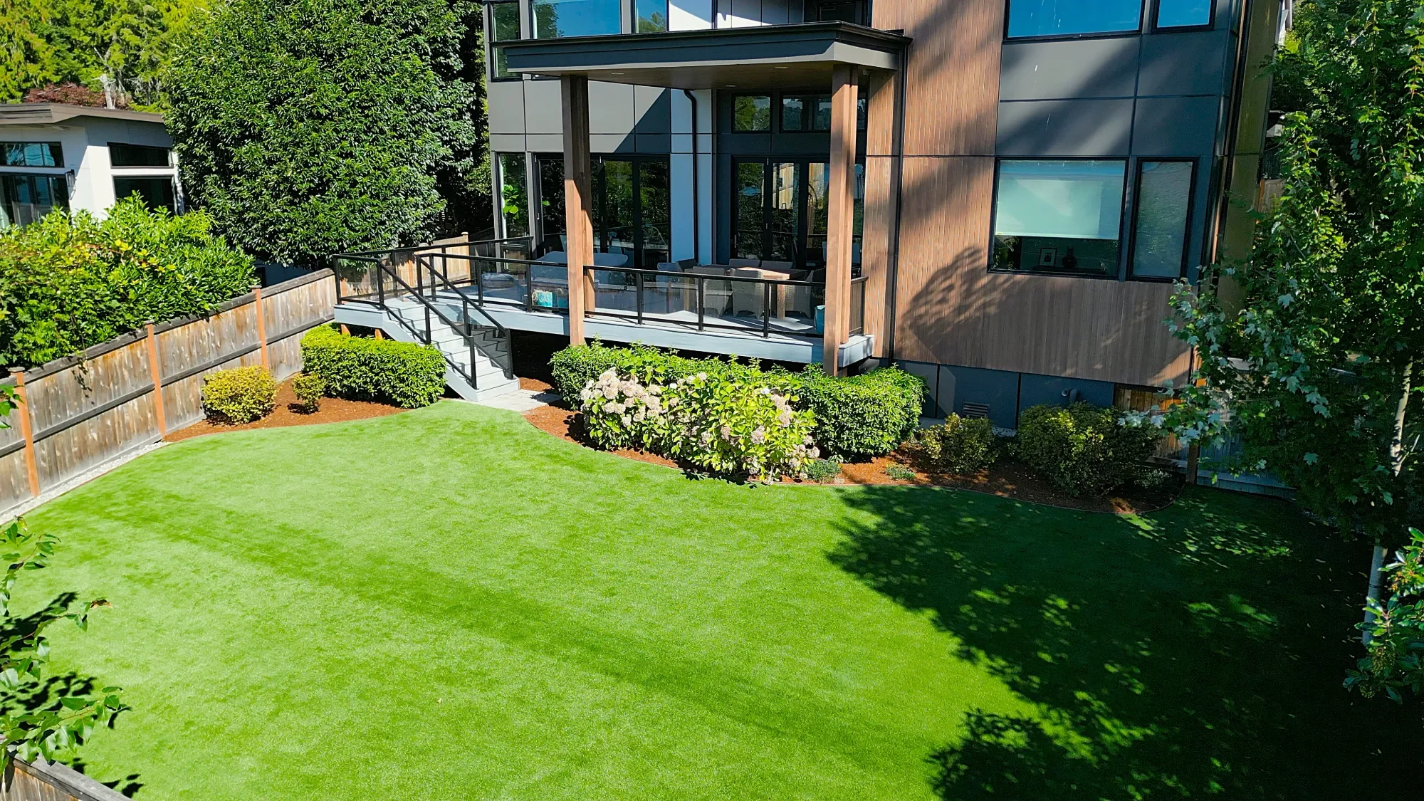 Modern backyard in Bothell with artificial turf installation, landscaped shrubs, and raised deck with glass railing