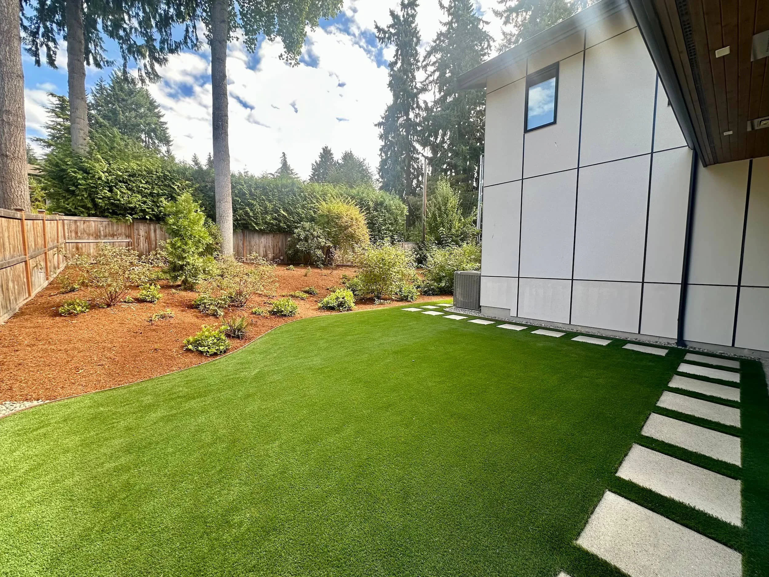 Modern backyard with green turf, stepping stones along the house, and a garden with small shrubs and trees, enclosed by a wooden fence, with clear blue sky and trees in the background.