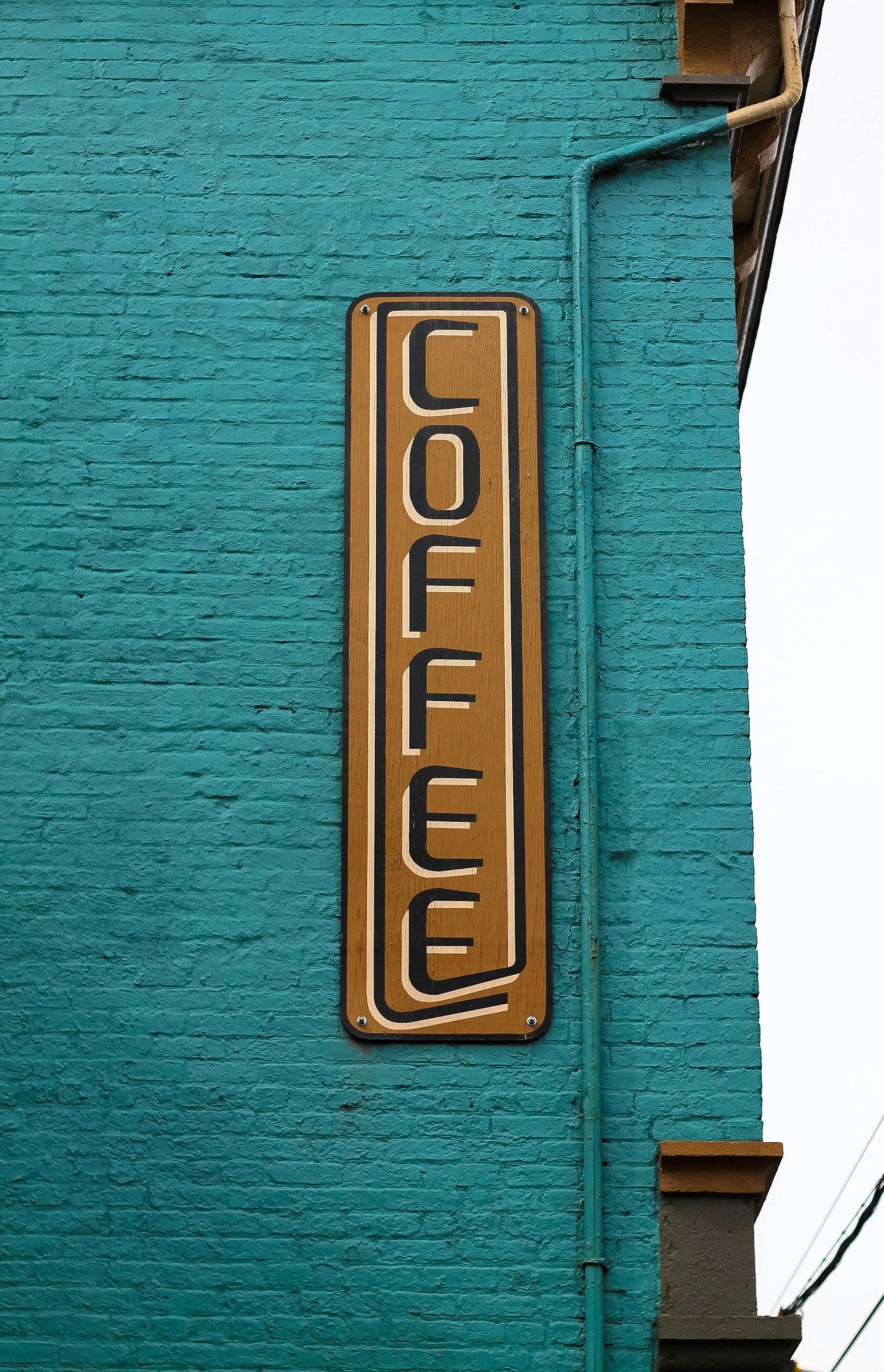 A vertical wooden sign with the word 'COFFEE' written in stylized black letters on a textured teal brick wall.