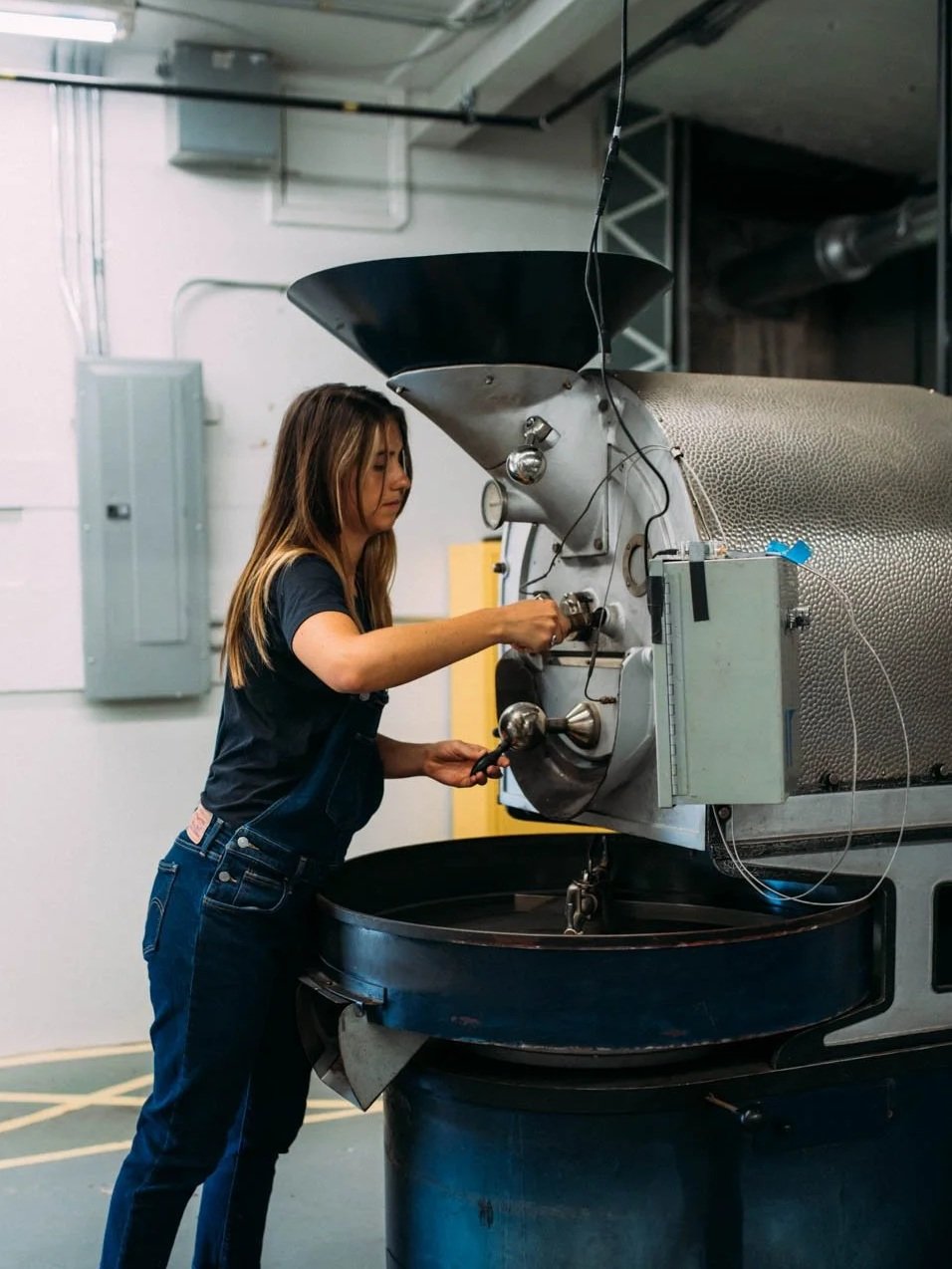 A woman operating a large industrial coffee roasting machine in a factory or warehouse setting.
