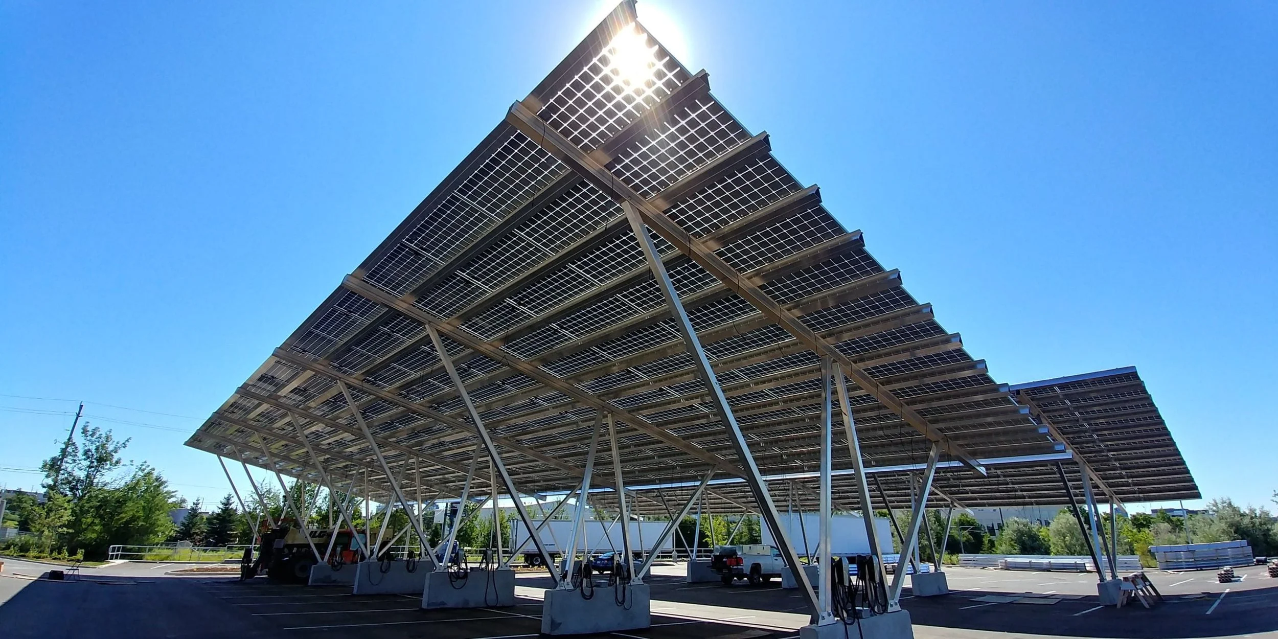 A large solar panel array installed in a parking lot, with several vehicles parked underneath and a bright blue sky overhead.