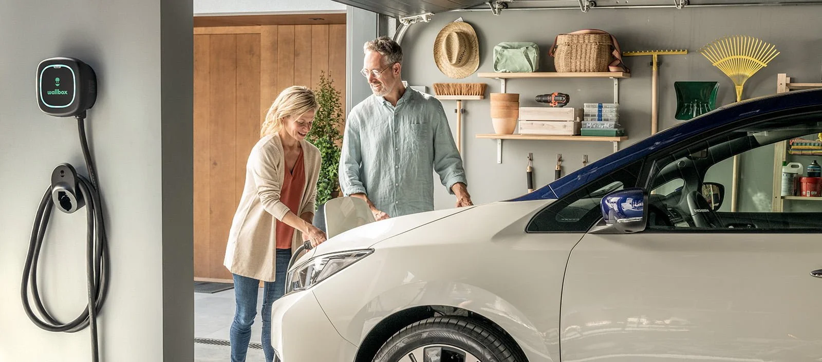 A man and woman loading items into the trunk of a white electric car in a garage, with an electric vehicle charging station on the wall.