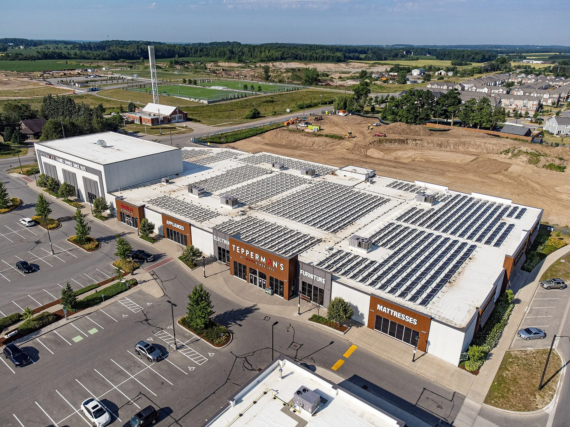VCT Group Rooftop Solar at Tepperman's Kitchener showroom. A large retail store with solar panels on its white roof, parking lot with cars, and landscaping, situated in a suburban area with construction and green fields in the background.