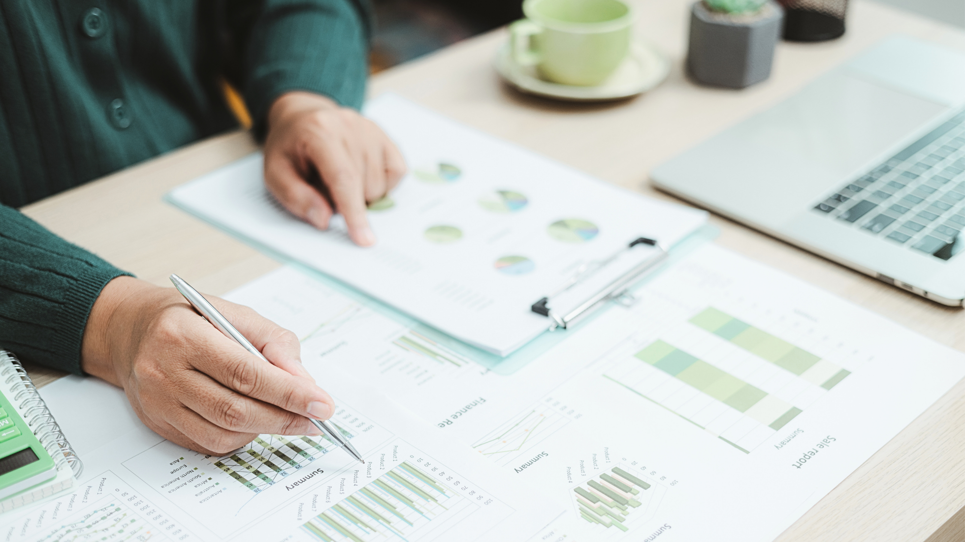 Close-up of a person's hands working on financial reports, with charts, graphs, a calculator, and a laptop on the table.