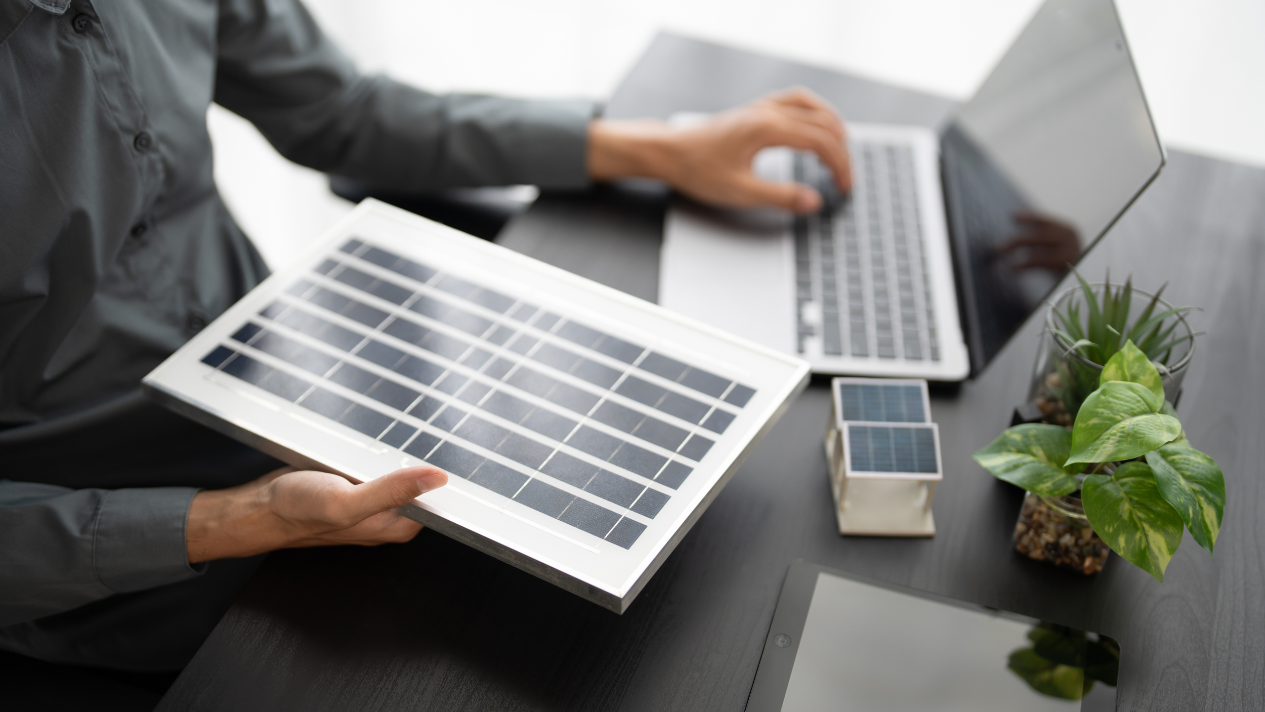 Person holding a solar panel sample at a desk with a laptop, miniature solar panel models, green plants, and a tablet.