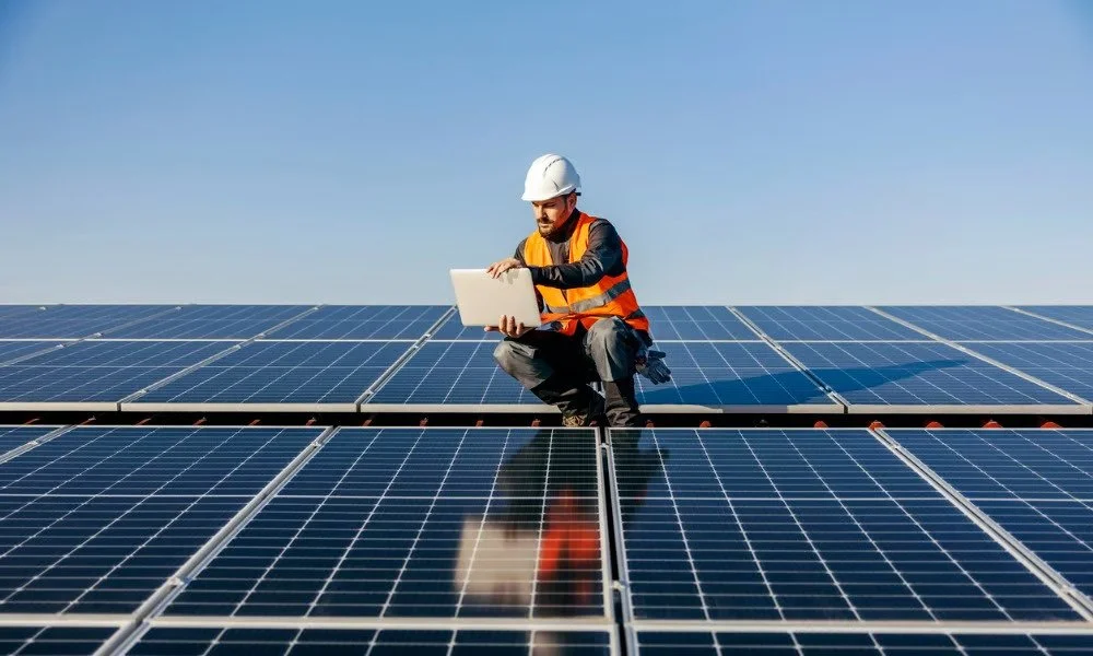 A man in a hard hat and safety vest kneeling on solar panels with a laptop in hand, outdoors under a clear sky.