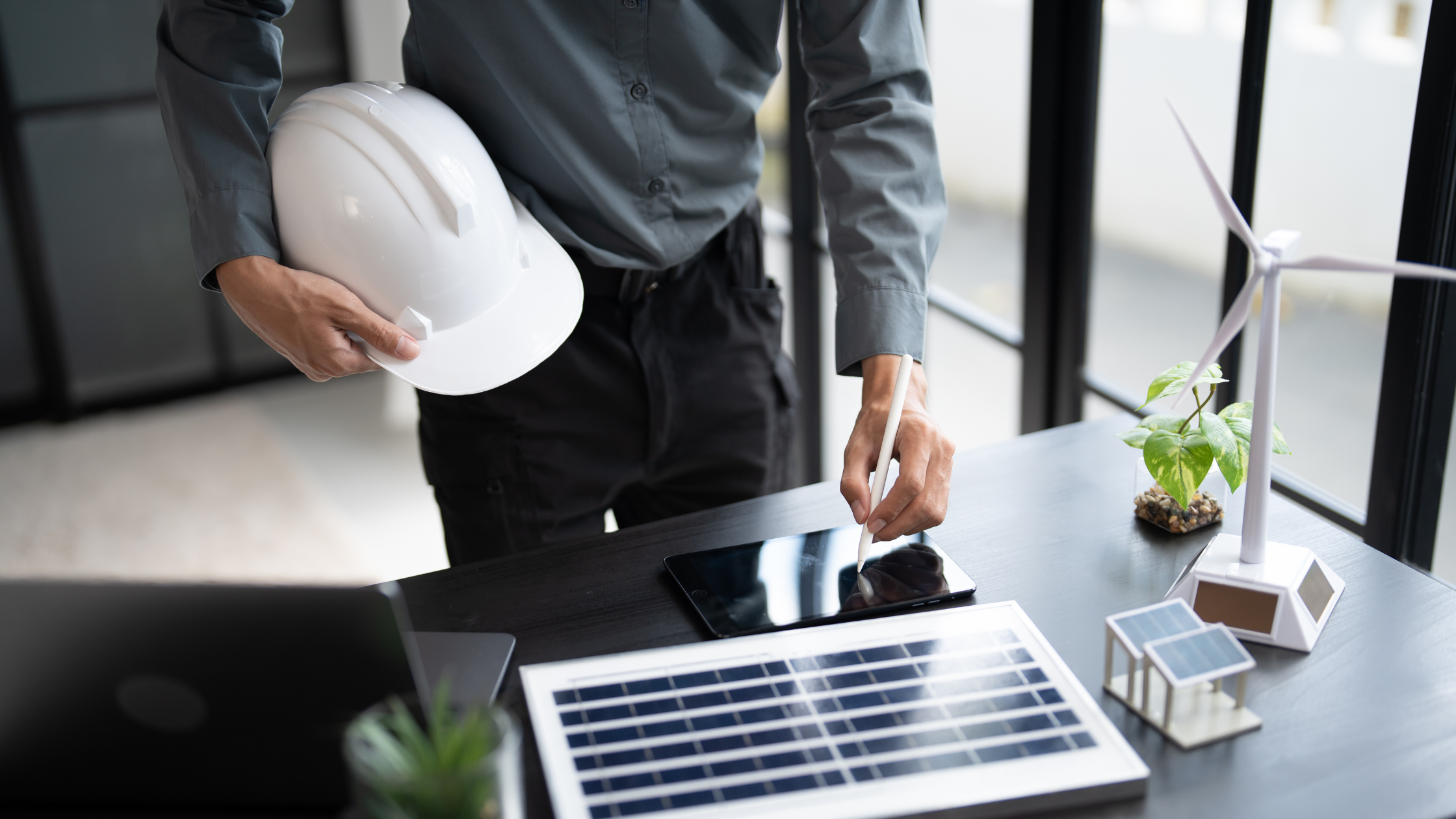 A person wearing a gray shirt holding a white safety helmet, working on a tablet with a stylus on a black table. The table has solar panels, a small wind turbine model, a plant, and miniature house models.