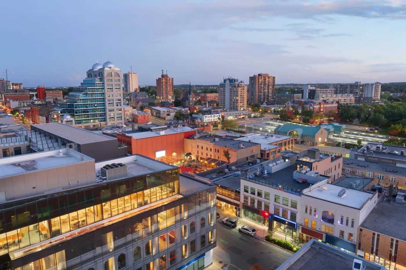 A cityscape view of Kitchener during dusk with illuminated buildings, streets, and a clear sky above, showcasing a mix of modern and historic architecture.