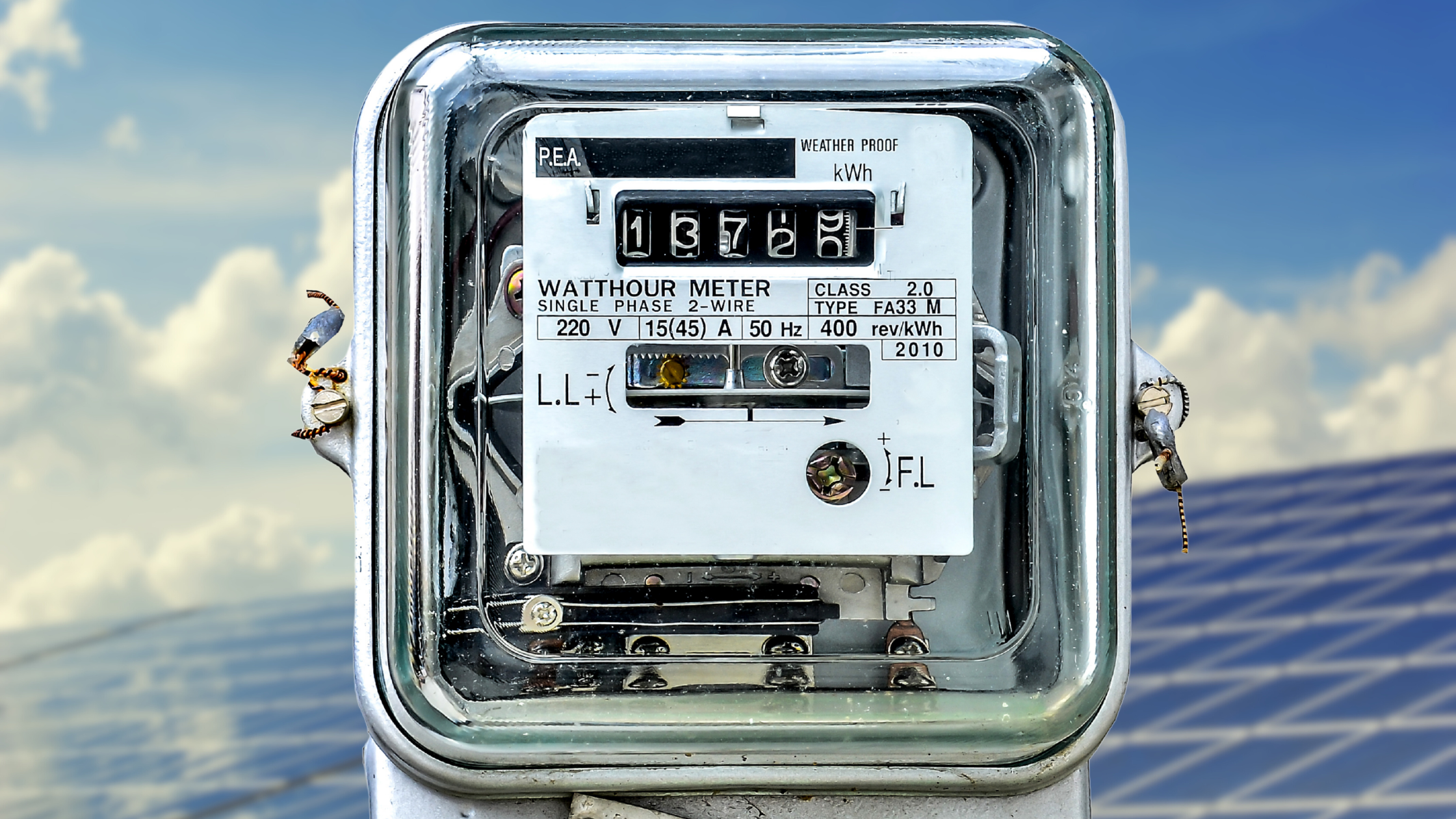 Close-up of an electric meter mounted outdoors with solar panels and a cloudy sky in the background.
