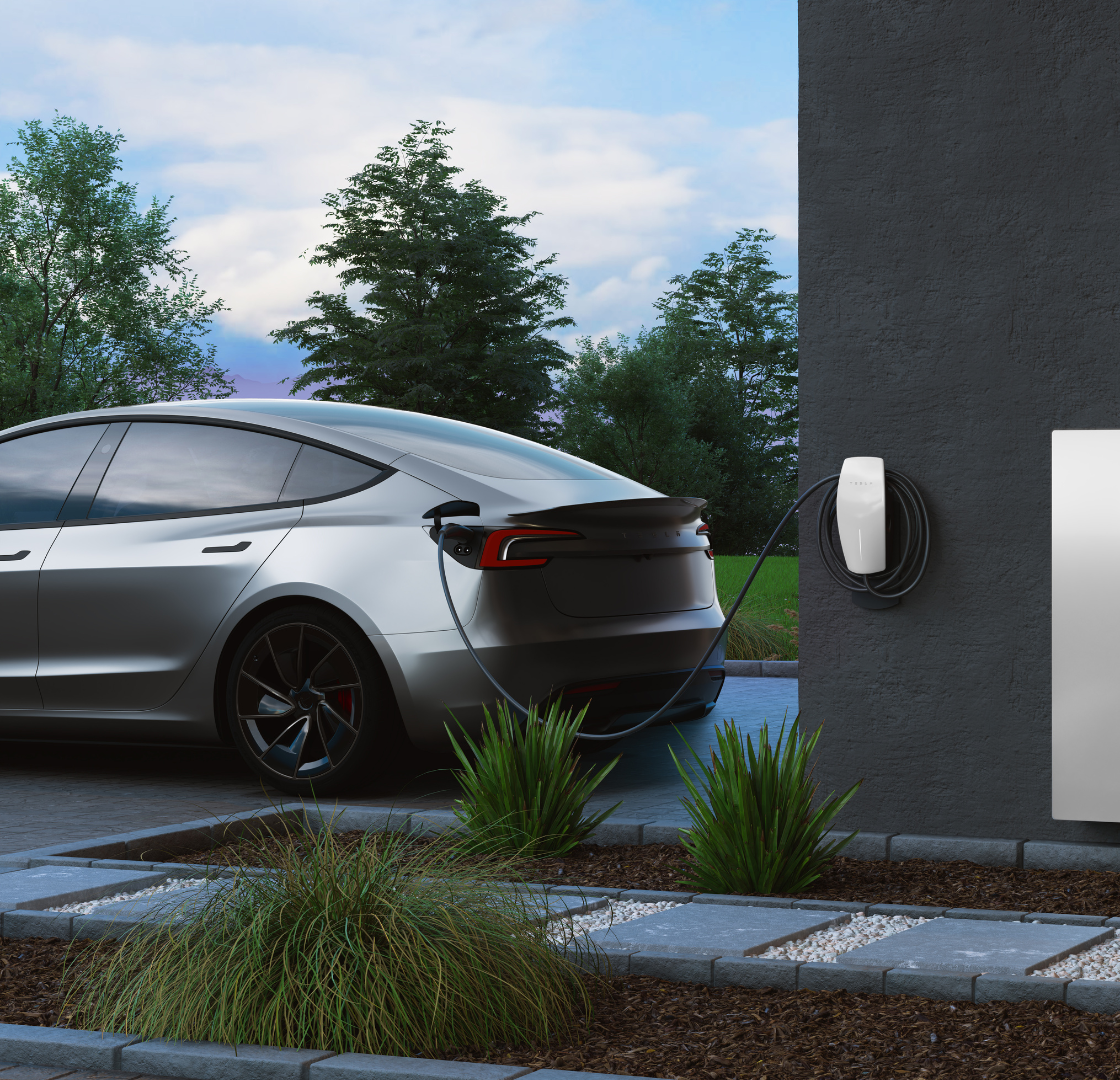 A silver electric car charging at a wall-mounted charging station outdoors with trees and blue sky in the background.