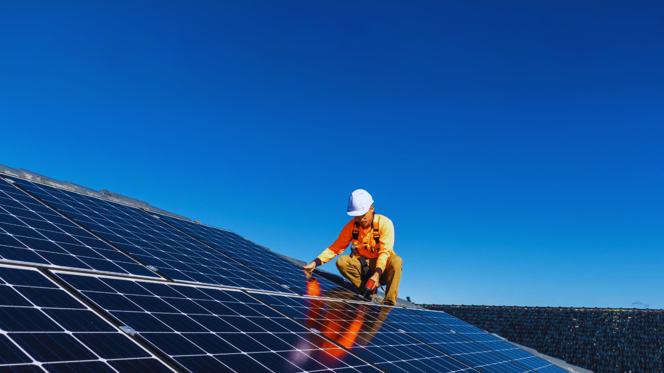 A worker wearing an orange shirt, beige pants, a white safety helmet, and a harness kneels on a large array of solar panels on a roof, adjusting the panels against a clear blue sky.
