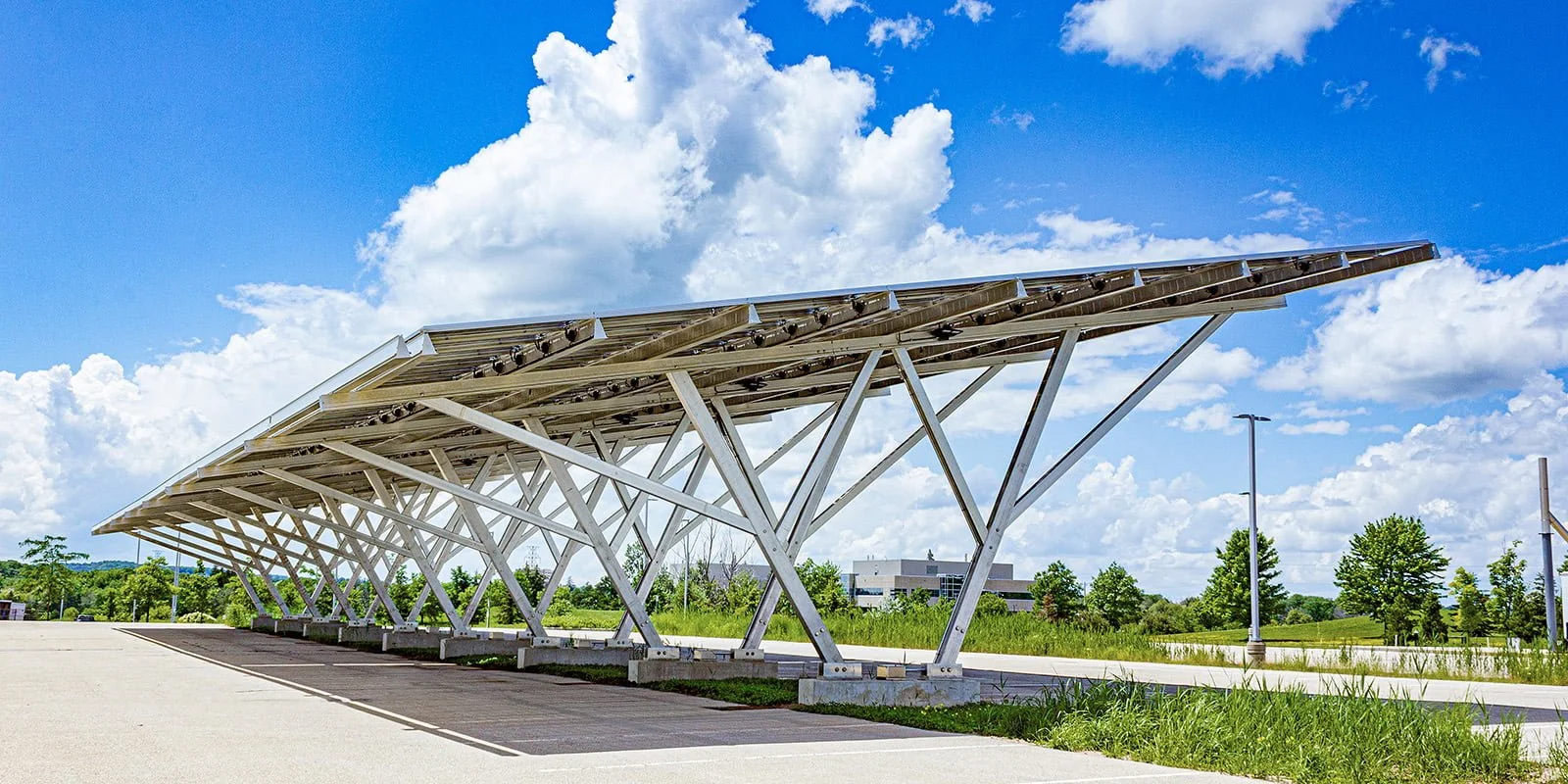 VCT Group solar carport system. Modern parking shelter with angled steel beams, open sides, under a partly cloudy sky with trees in the background.