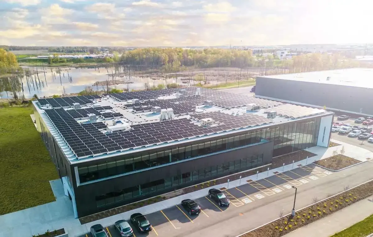 An aerial view of a modern office building with a rooftop covered in solar panels, parking lot in front, and a water body with trees nearby.