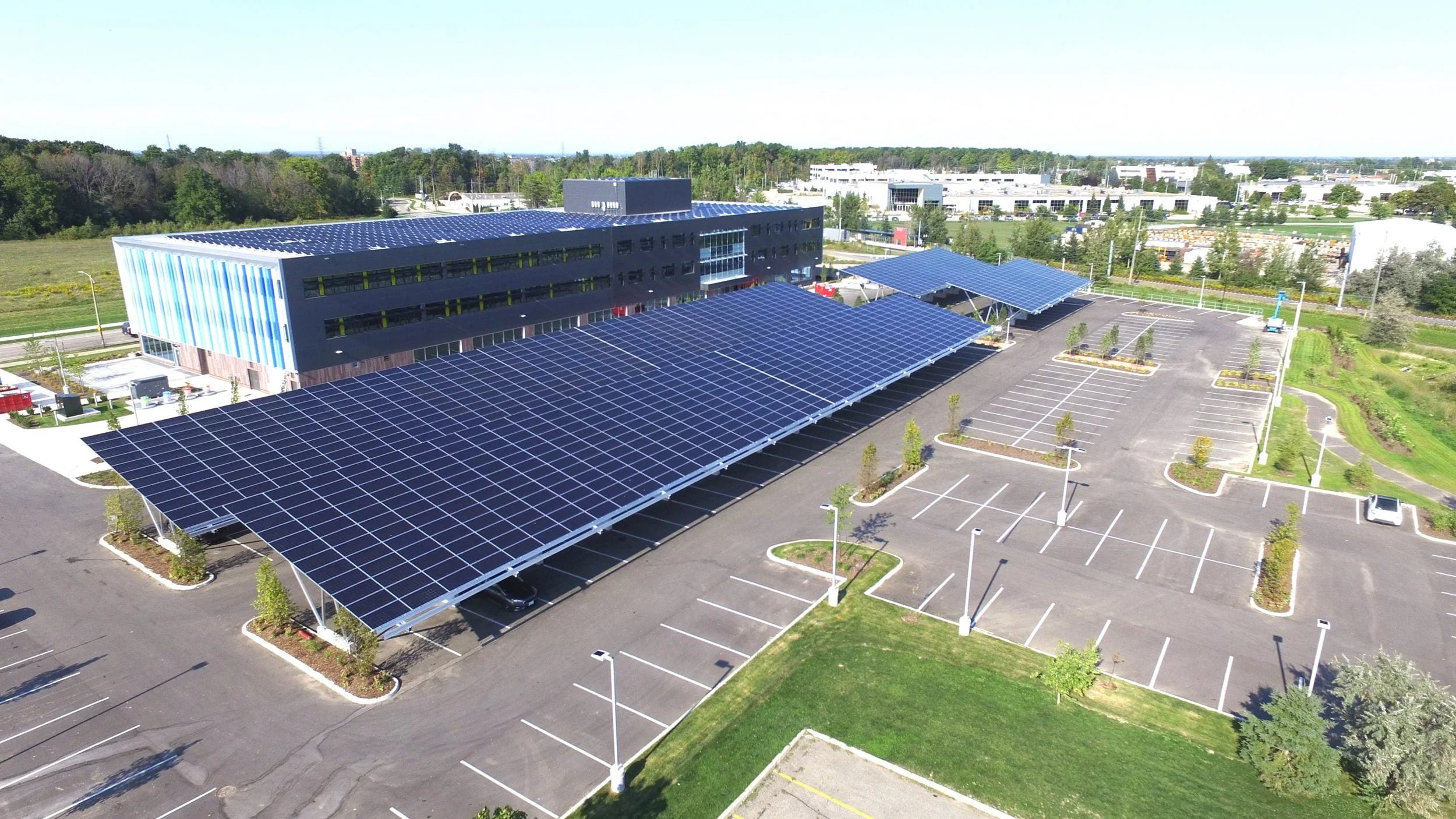 VCT Group solar carport system and rooftop solar panels in Ontario, Canada.A building with solar panels on the roof, surrounded by an empty parking lot with few cars, trees, and a distant view of a cityscape.