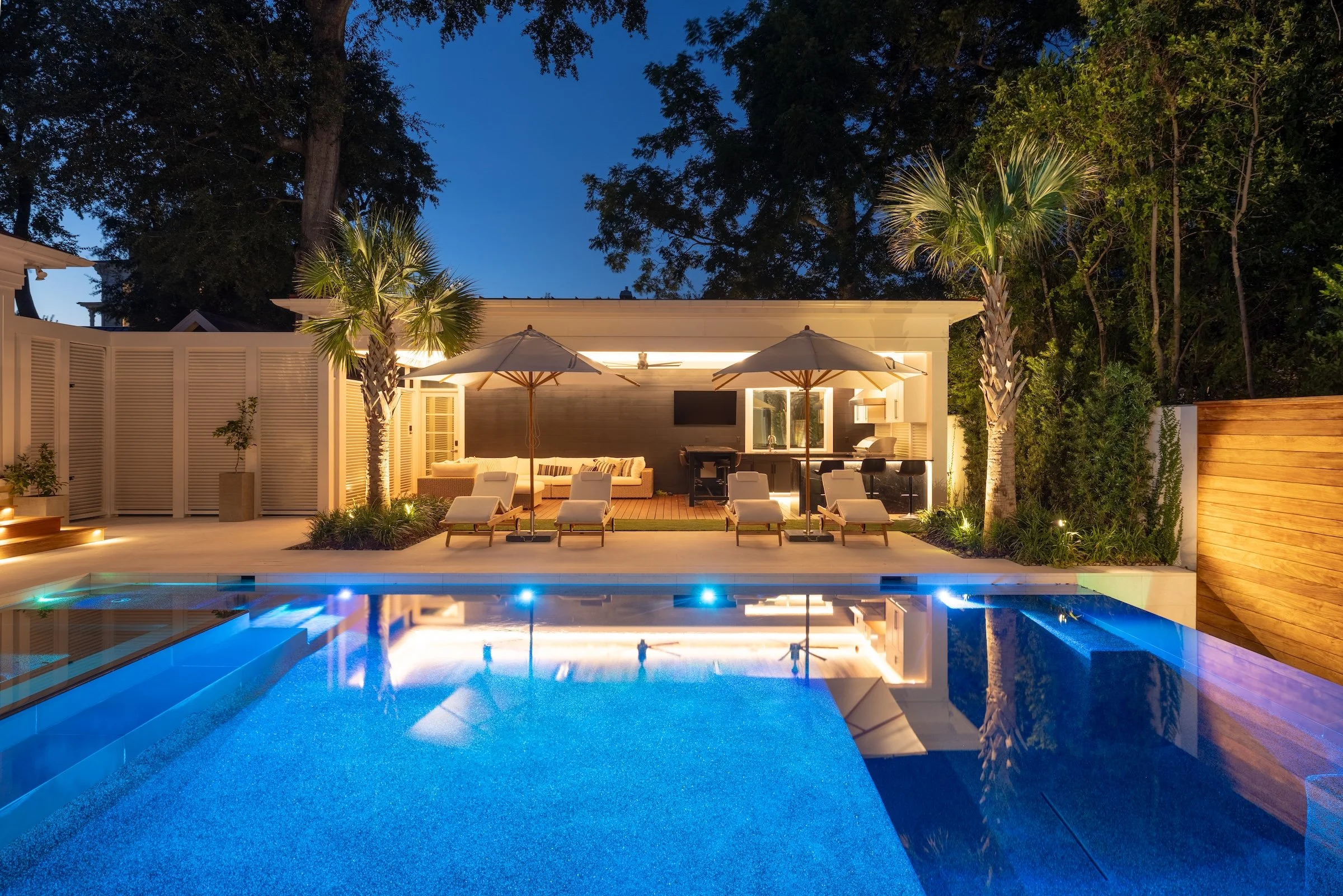Inground pool lit up at night with pool loungers, paver patio and outdoor living space behind it