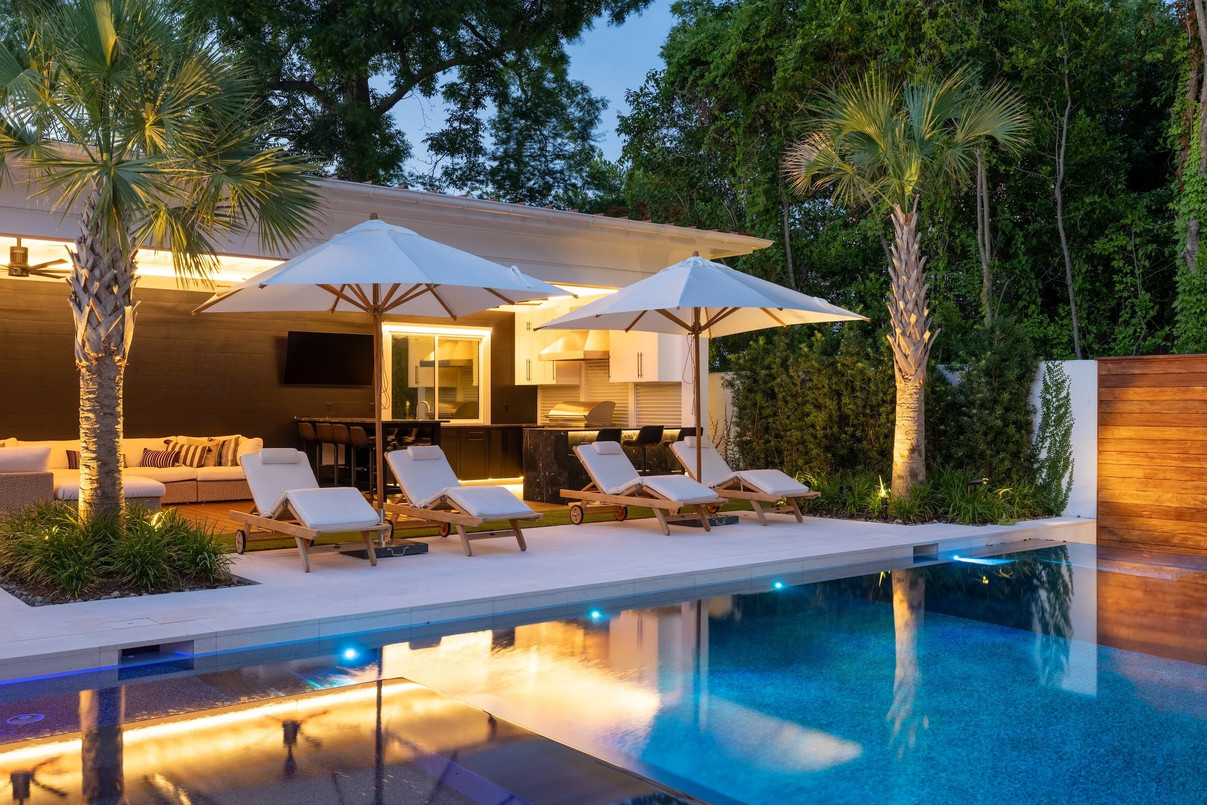 inground pool with lights on, paver patio, and outdoor kitchen at night