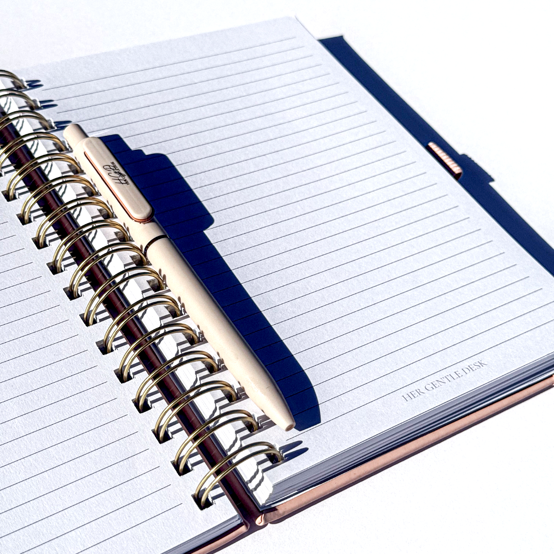 Open spiral-bound notebook with lined pages, a white pen resting on the pages, and a second closed notebook underneath. Sunlight casting shadows on the pages.