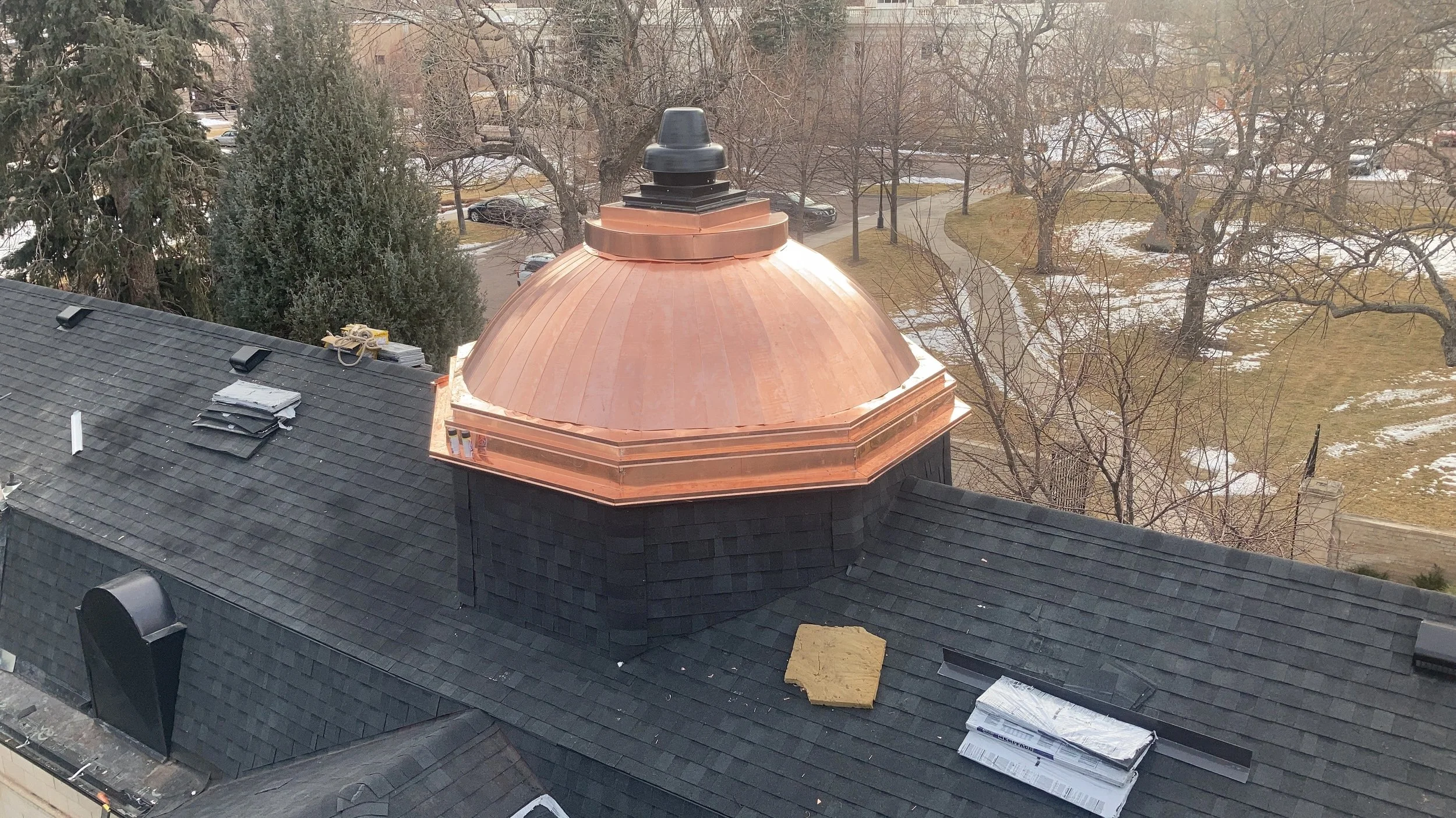 An aerial view of a roof with black shingles, featuring a large copper dome structure at the center, and several other vents and materials on the roof. In the background, there are leafless trees, parking lot, and patches of snow on the ground.