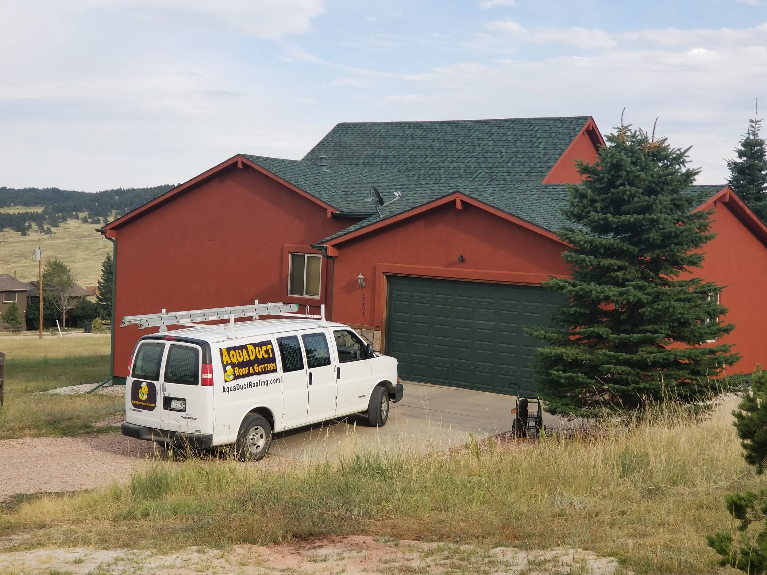 A red house with green roof and garage, surrounded by grass and trees, with a white van parked in the driveway advertising AquaDuct roofing services, and a ladder mounted on top of the van.