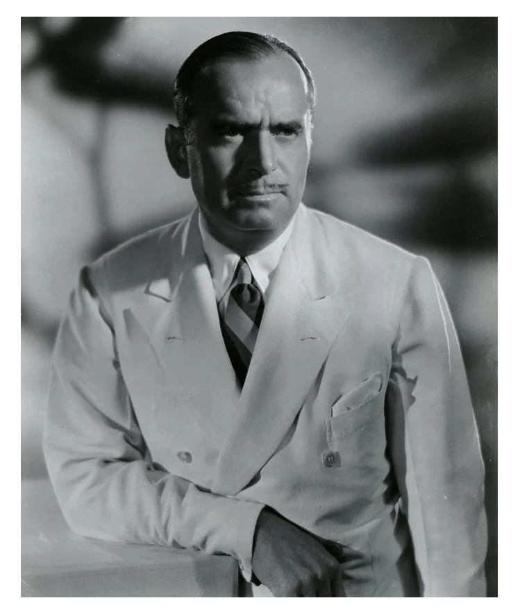 Black and white photograph of a serious man in a light-colored suit with a striped tie, leaning on a surface, with a blurred background.