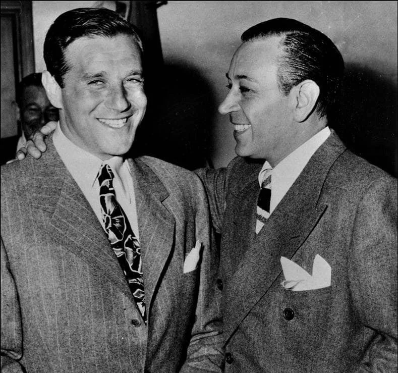 Two men in suits smiling and greeting each other at a social event, with one placing his hand on the other's shoulder.