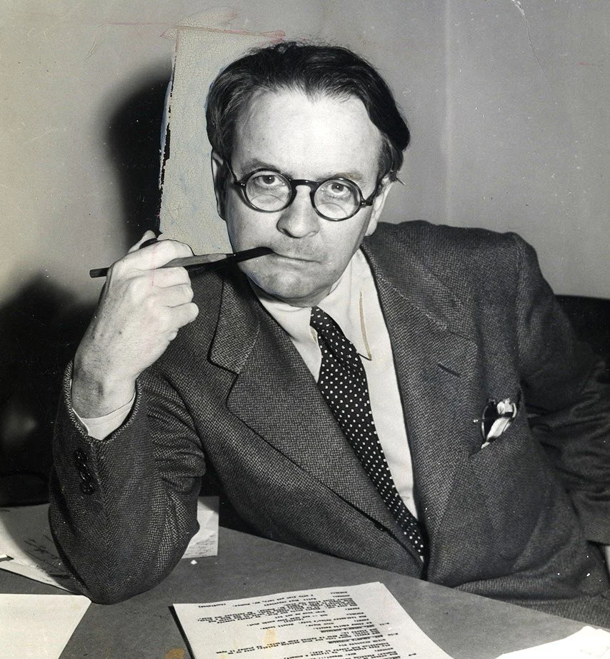 Black and white photo of a man with glasses sitting at a desk, holding a pipe near his mouth, with papers on the desk.