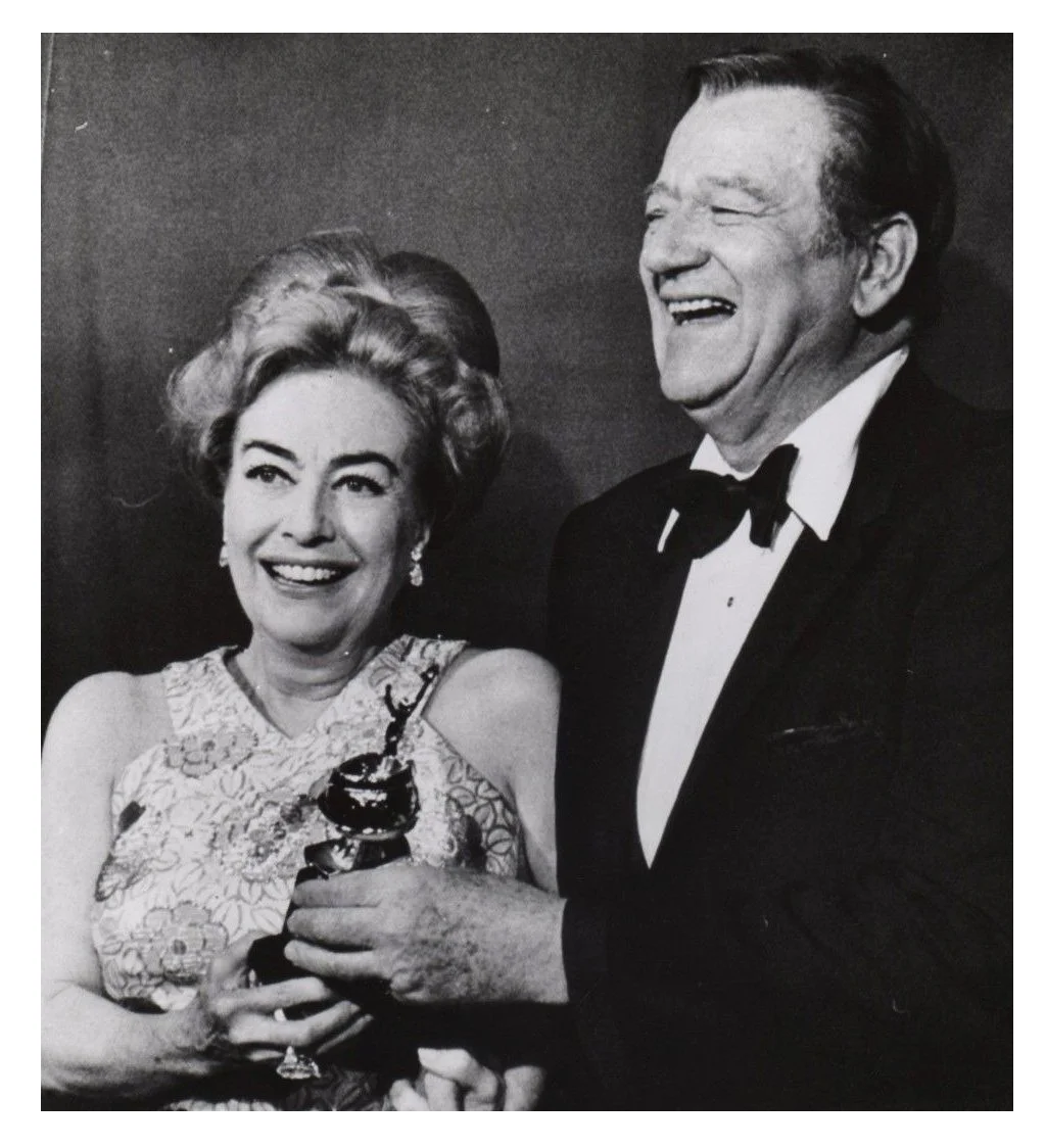 Black and white photo of a woman holding an award statue and a man wearing a tuxedo, both smiling and looking happy.