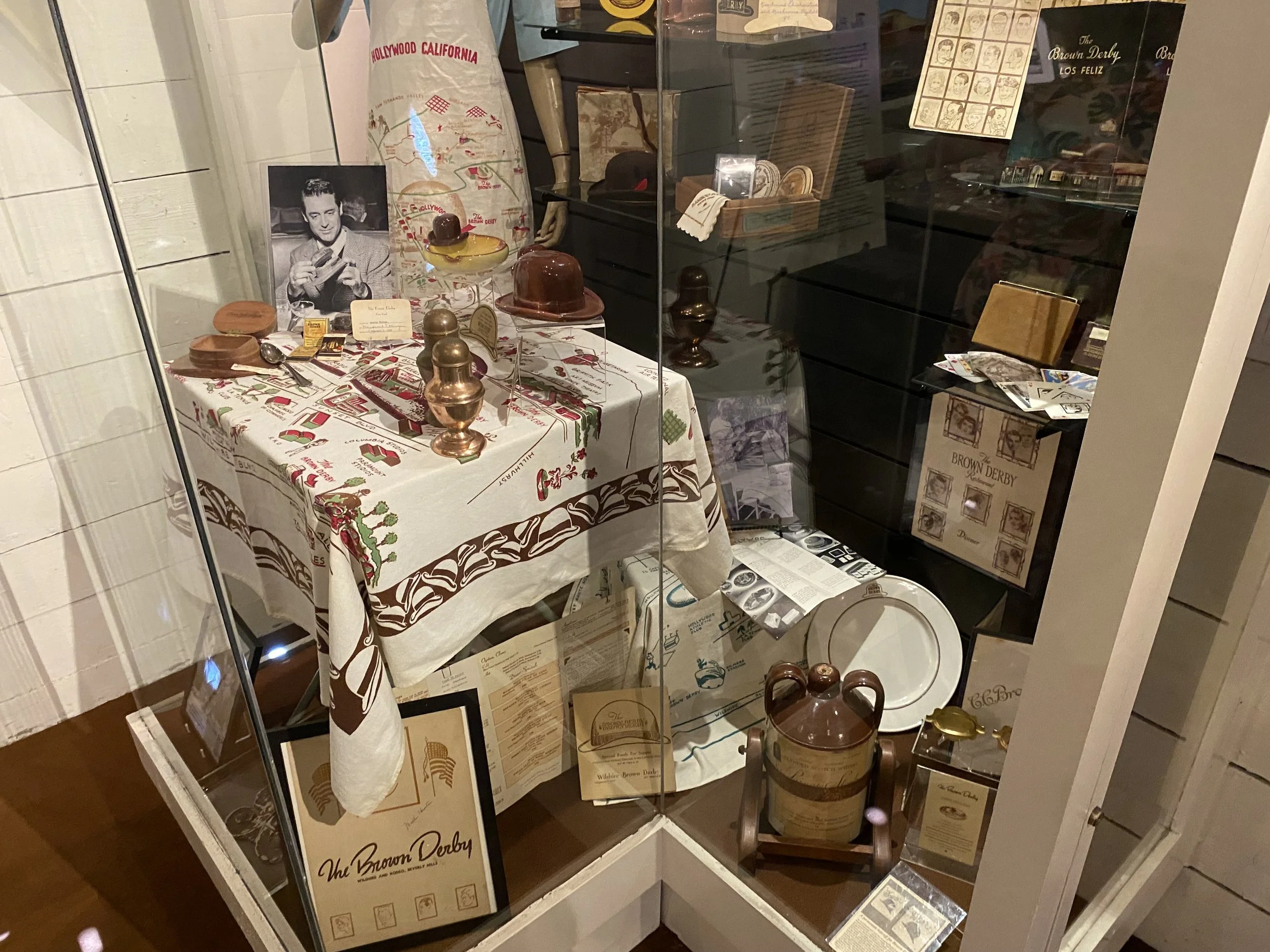 Display of vintage items including a black-and-white photograph of a man, brass and wooden decorative pieces, a ceramic pitcher, and various memorabilia on a table and shelves in a museum or exhibit setting.