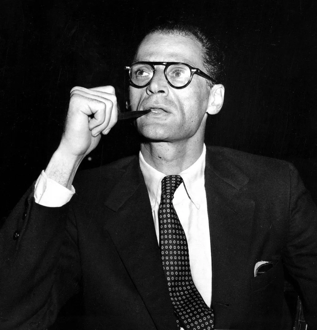 Black and white photograph of a man in a suit, wearing glasses, holding a pipe in his mouth and looking off to the side.