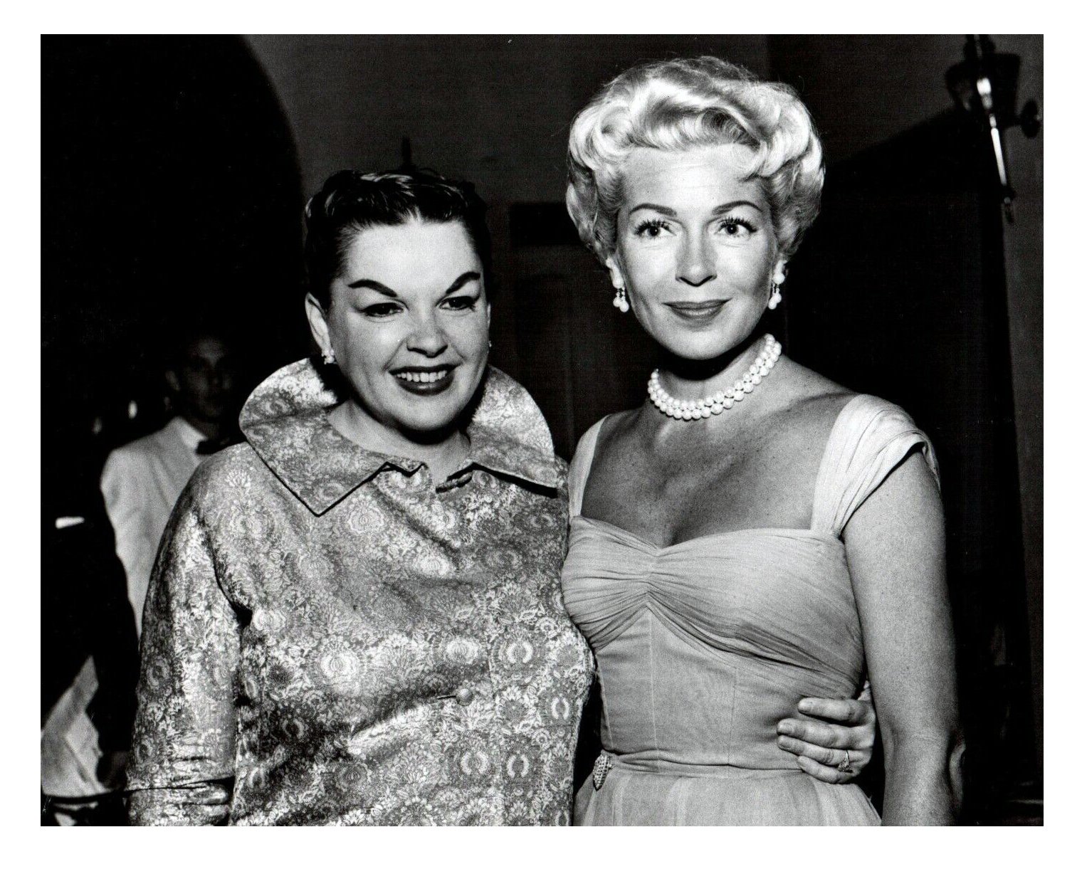Black and white photo of two women smiling, dressed in vintage style, at a social event, with a person in a white suit in the background.
