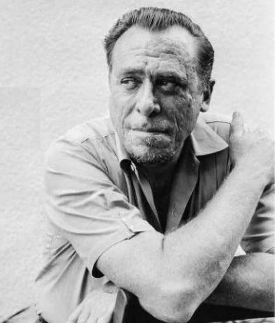 Black and white photo of a man with a disfigured face, looking to the side, wearing a collared shirt with rolled-up sleeves, sitting with his arms crossed.