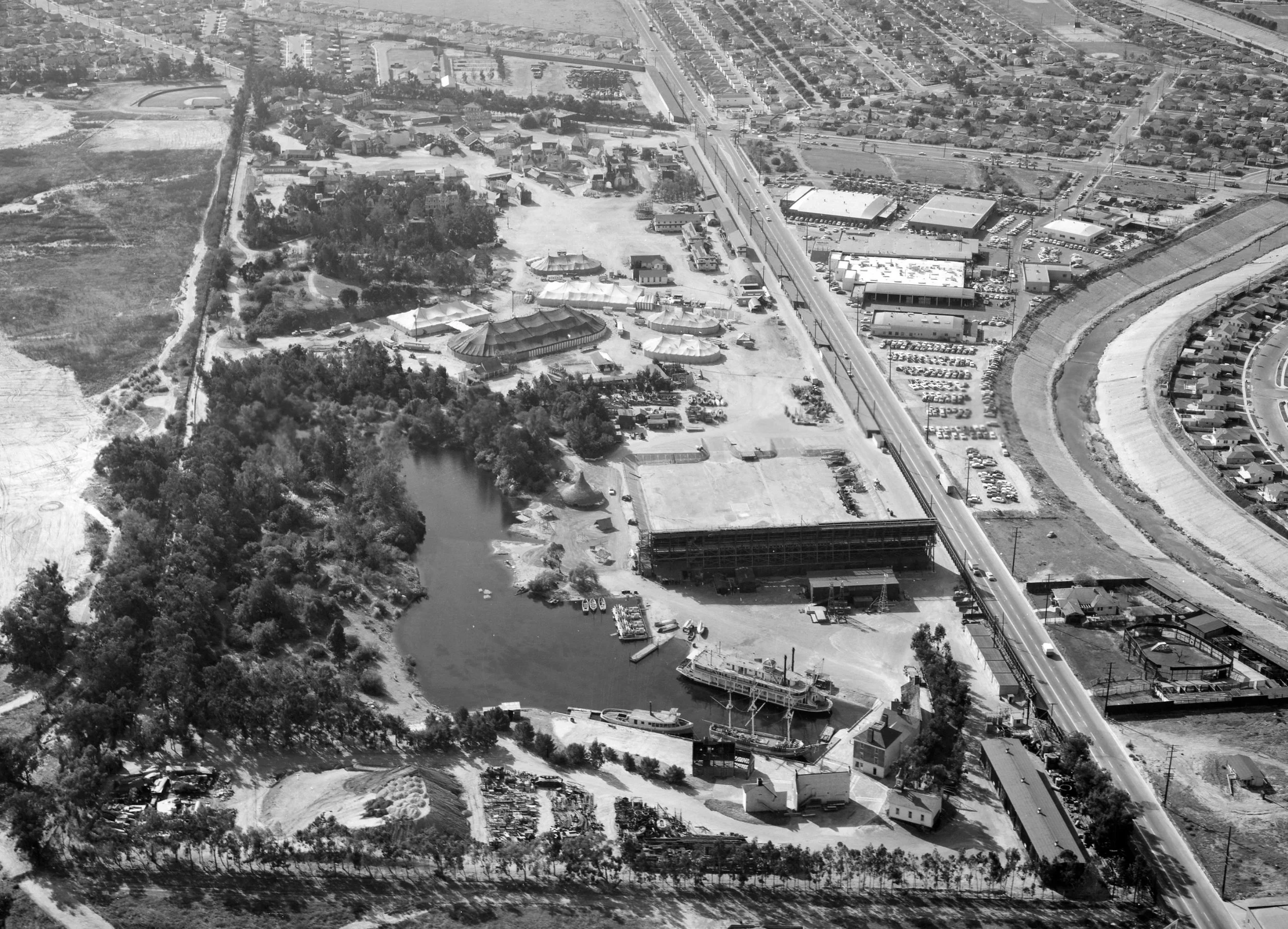 Aerial view of a park with a lake, surrounded by trees, amusement park rides, and a gravel area. Nearby are roads, parking lots, commercial buildings, and residential neighborhoods.