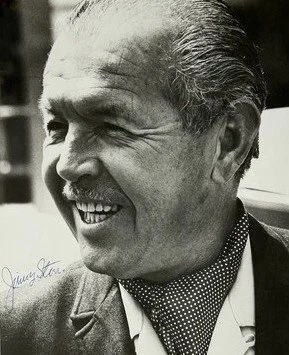 A smiling elderly man with slicked-back hair, wearing a suit jacket and a polka dot scarf.
