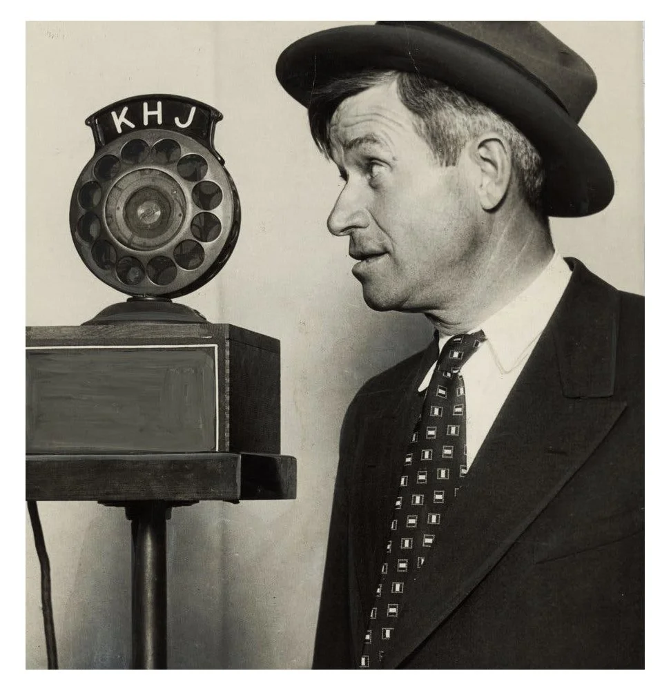 A black and white photo of a man wearing a hat and suit, looking at an old-fashioned rotary telephone with the letters 'K H J' on it, sitting on a wooden stand.