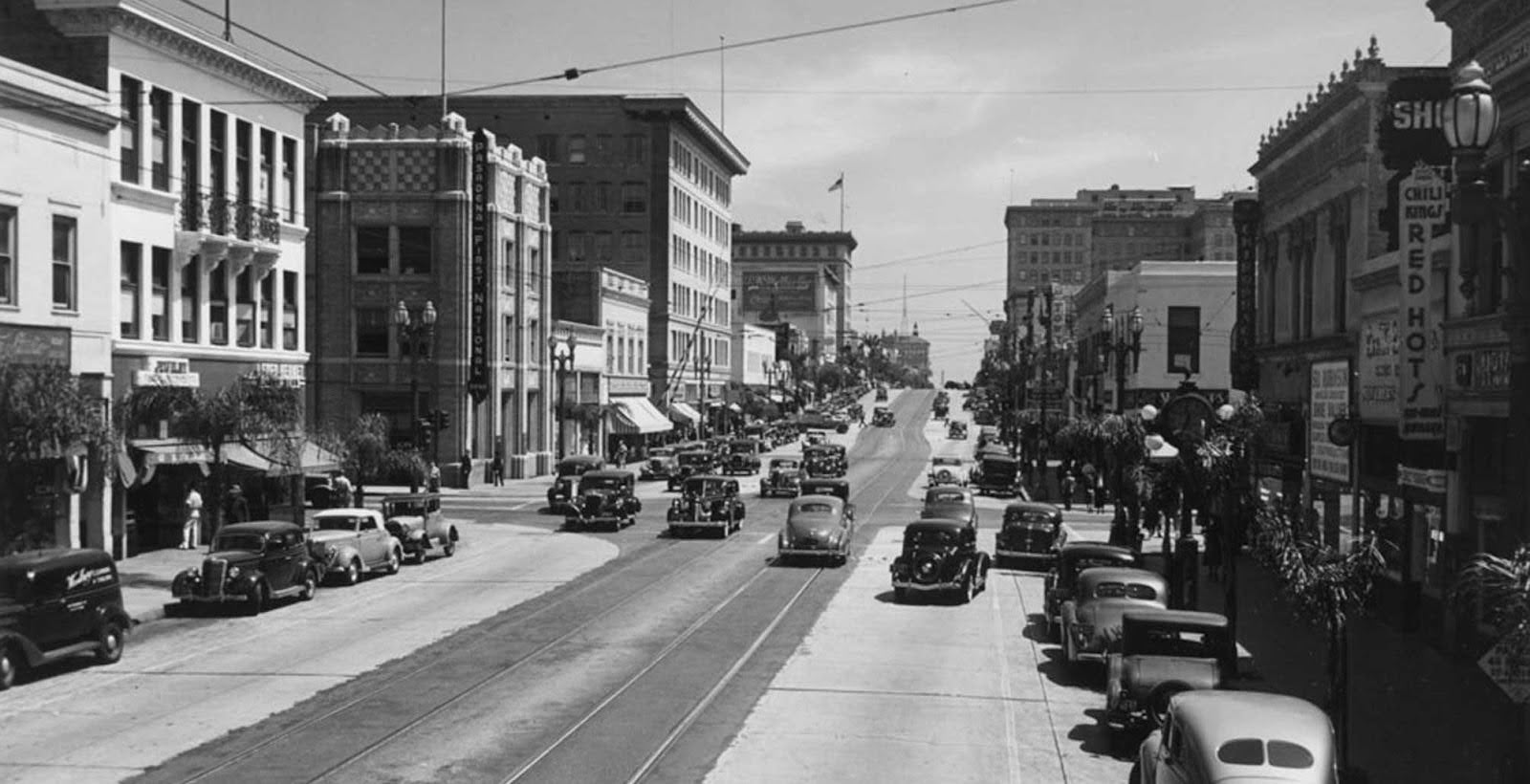 Black and white photo of a busy downtown street with vintage cars parked along the sides and moving down the street. Multi-story buildings line both sides of the street, some with signs and awnings. Utility lines run overhead, and a hill slopes upward in the background.