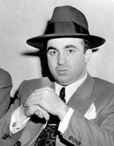 Black and white photo of a man wearing a fedora hat, suit, and tie, with a serious expression, sitting with hands clasped.