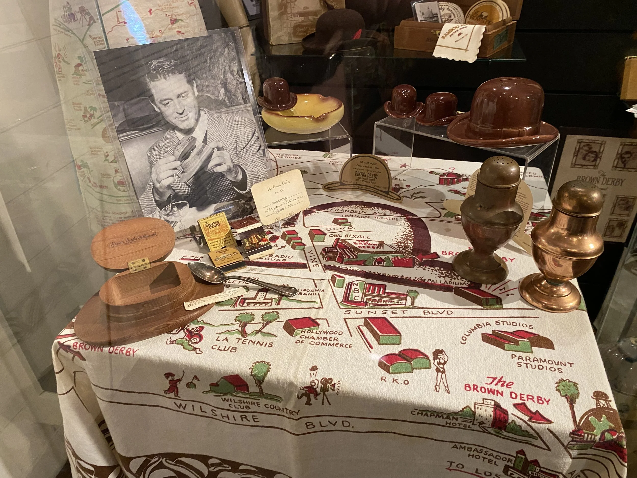 Display of vintage items including metal and wooden trophies, a black-and-white photograph of a man holding a sandwich, and a cloth with a colorful map of Brown Derby and Hollywood area landmarks.