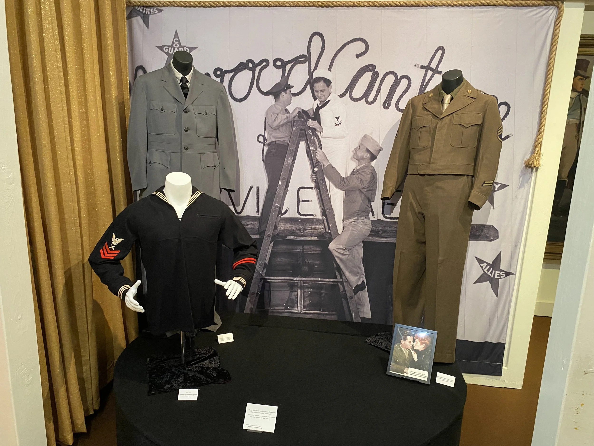 Display of vintage military uniforms including a black sailor uniform with red chevron patches, a gray officer's uniform, a brown military jacket with pants, and a beige military uniform with pants, arranged in front of a historic black-and-white pho