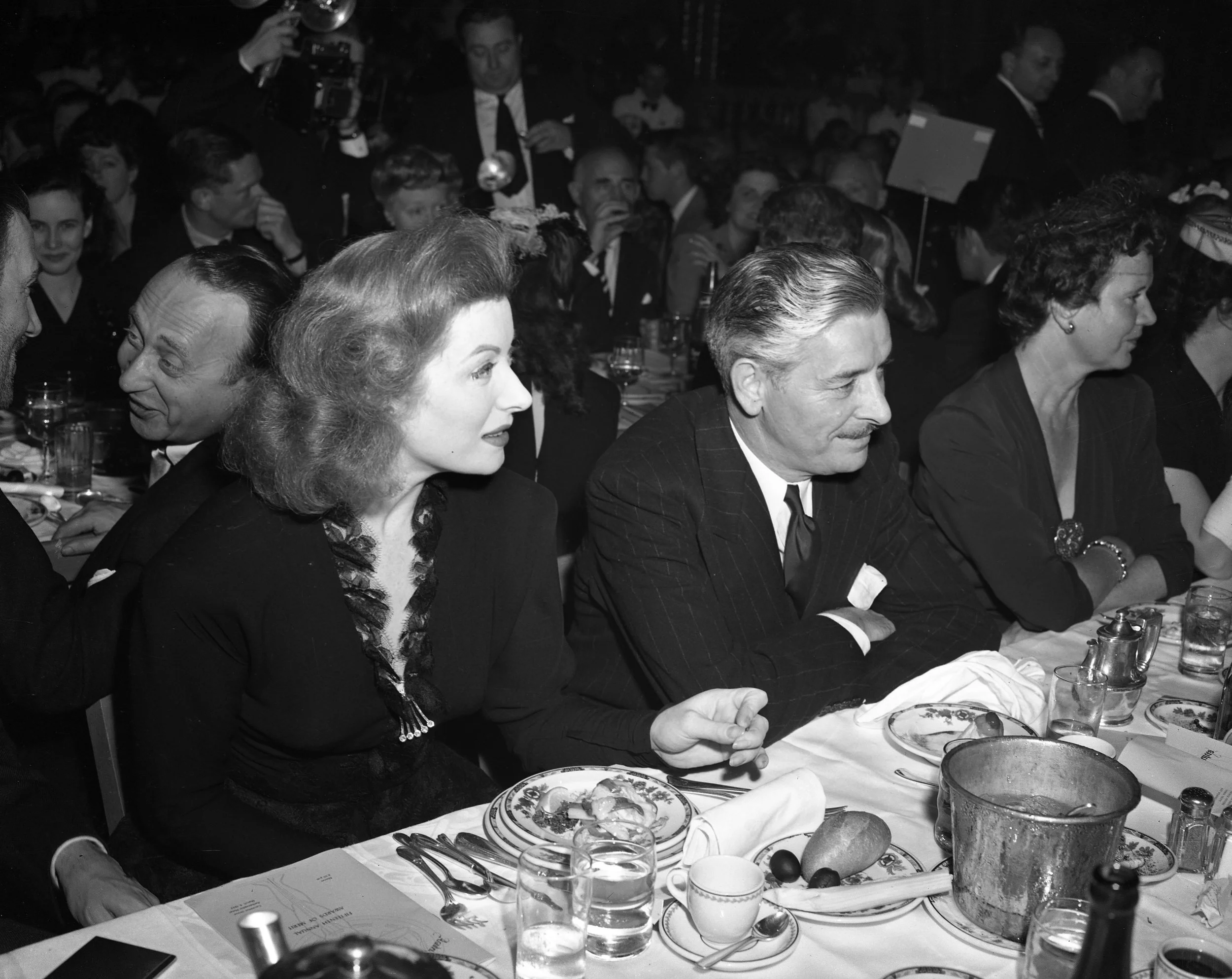 People sitting at a table during a formal event or dinner. The table is set with plates, glasses, utensils, and food items. The background shows more people and some standing, all dressed in formal attire.
