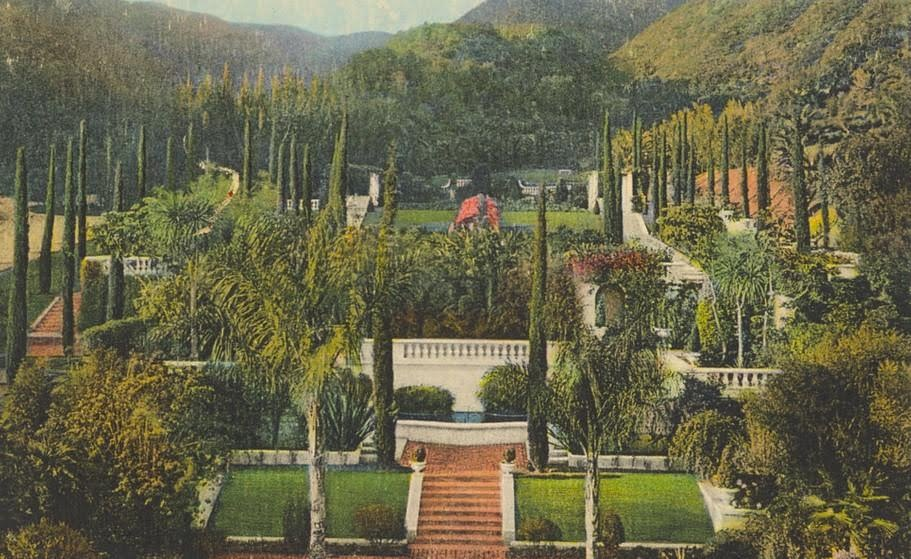 A scenic landscape featuring a large, beautifully landscaped garden with steps leading to a white building with arches, surrounded by tall cypress trees and lush greenery, with mountains in the background.